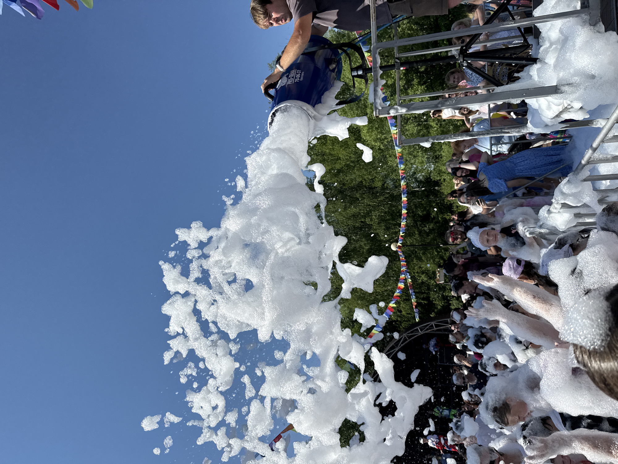 In Заволжье, Russia, a foam party is underway. A large blue foam cannon, operated by a man wearing a grey t-shirt, is spraying a torrent of white foam into the air. The foam is cascading down onto a crowd of people who are enthusiastically interacting with the foamy deluge. Many individuals are visible with their arms raised, some appearing to be touching or playing in the foam. The background features trees and a bright blue sky, with colorful triangular flags strung across the scene, suggesting a festive outdoor event. The overall impression is one of fun and celebration in a public outdoor setting.