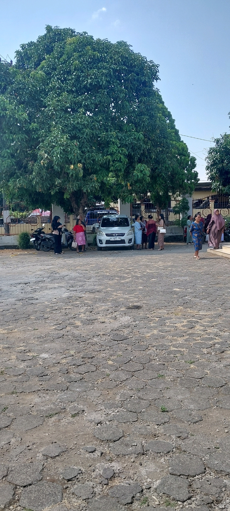 A group of people are gathered under the shade of a large tree next to a white car and a motorcycle in Keling, Indonesia. The ground is paved with hexagonal interlocking stones, which are cracked and worn in many places. The scene appears to be a public area, possibly a parking lot or a gathering space. The people are dressed in casual and traditional clothing, with some women wearing headscarves. The sky is clear and blue.