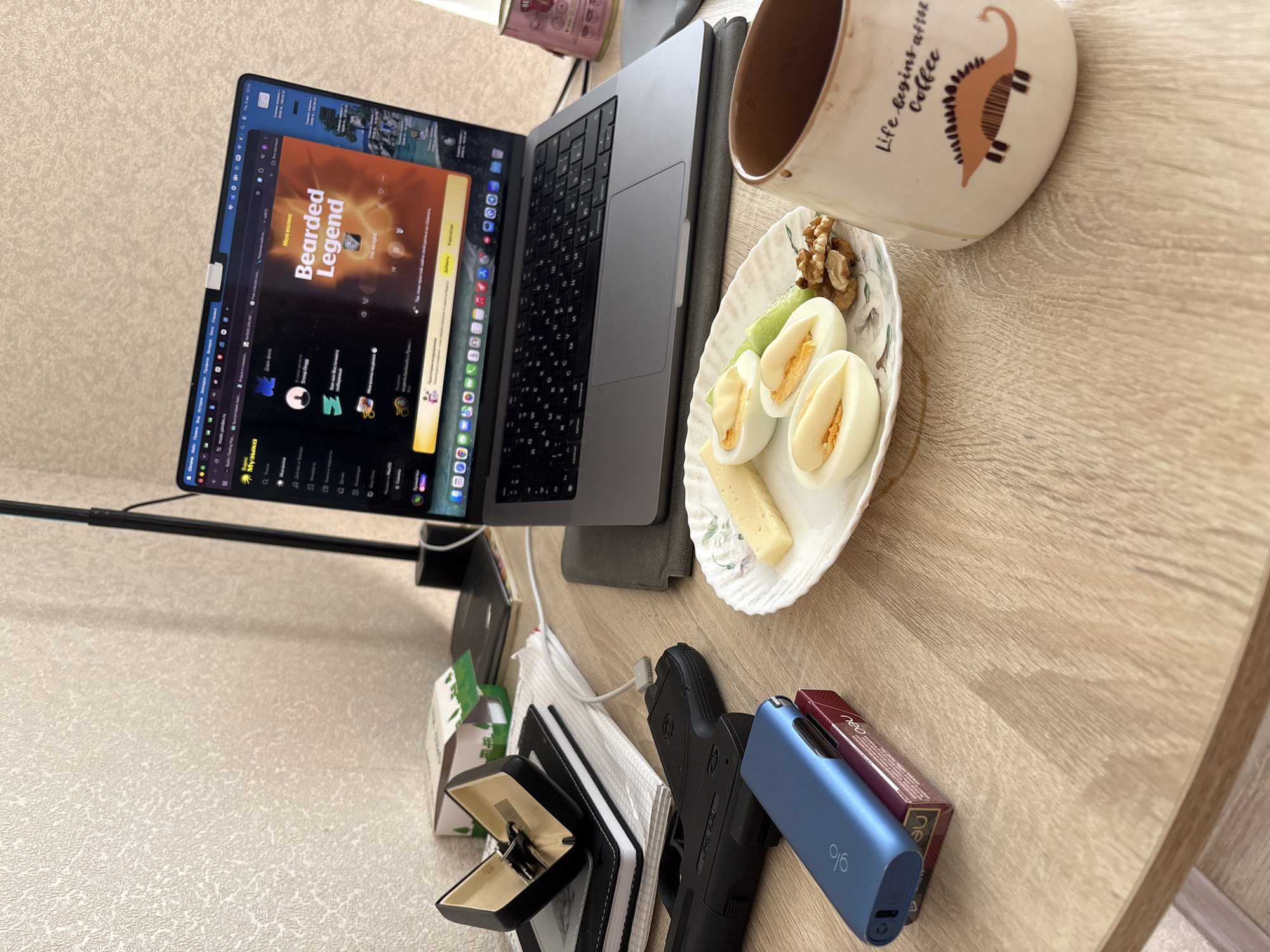 The image depicts a close-up, eye-level, indoor scene of a wooden desk cluttered with various objects. On the left, a black handgun is placed next to a blue electronic smoking device resting on a pack of cigarettes. To the left of these items, a black and gold key holder containing keys sits open on top of a stack of notebooks and papers. A laptop, displaying a music streaming service with the title "Bearded Legend," is positioned in the center of the desk, angled towards the viewer. In front of the laptop, a white plate holds a breakfast assortment of hard-boiled eggs, sliced cheese, cucumber, and walnuts. To the right of the plate, a ceramic mug with a dinosaur illustration and the text "Life begins after Coffee" is visible. The background shows a textured, light-colored wall. The overall impression is that of a personal workspace with a mix of work, leisure, and personal items. The image does not contain any information that can be used to determine the specific location within the city of Saratov, Russia.