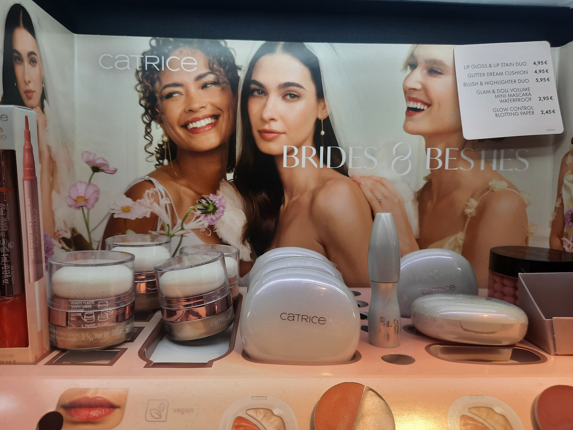 A retail display for Catrice cosmetics features a large promotional image of three smiling women, two of whom are wearing wedding veils, suggesting a bridal theme. The advertisement text reads "BRIDES & BESTIES". Below this, various Catrice beauty products are arranged, including what appear to be powder compacts, lip gloss, and mascara. A price list is attached to the display, listing items such as "LIP GLOSS & LIP STAIN DUO" for 4,95€, "GLITTER DREAM CUSHION" for 4,95€, "BLUSH & HIGHLIGHTER DUO" for 5,95€, "GLAM & DOLL VOLUME MINI MASCARA WATERPROOF" for 2,95€, and "GLOW CONTROL BLOTTING PAPER" for 2,45€. The display is well-lit and appears to be situated in a store environment. The overall aesthetic is clean and professional, typical of beauty product retail displays. The image was taken in Rosenheim, Germany.