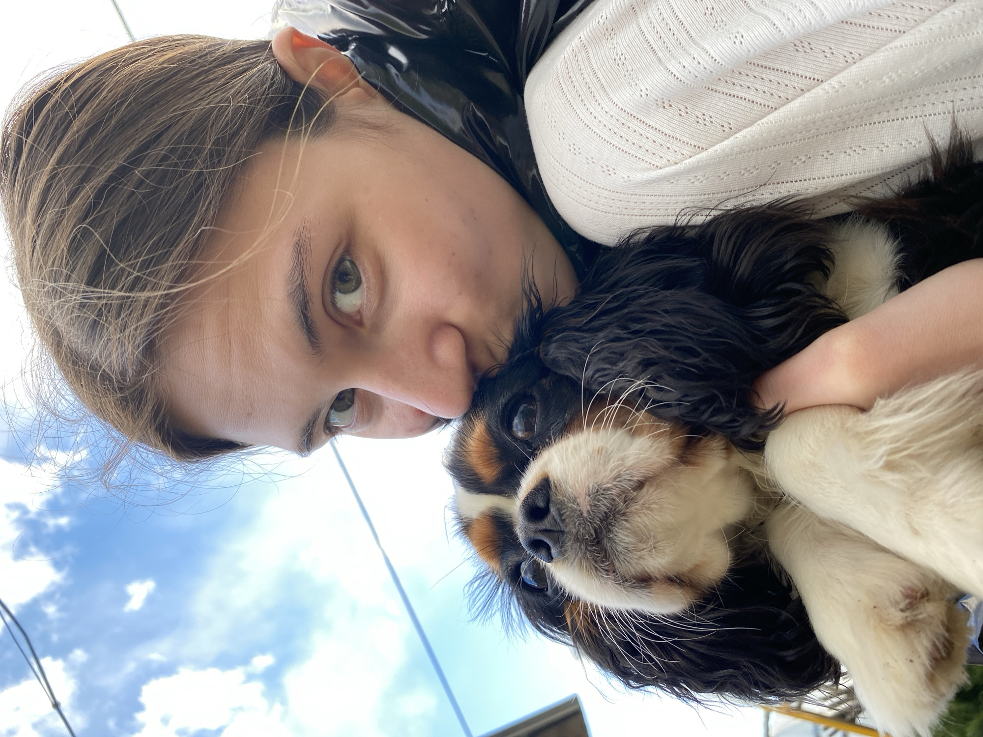 A young, fair-skinned person with light brown hair is holding a Cavalier King Charles Spaniel dog. The person is wearing a white top with a textured pattern and a dark, shiny jacket. The dog has black, white, and tan fur. The sky with clouds is visible in the background. The location is possibly the city of Votkinsk, Russia.