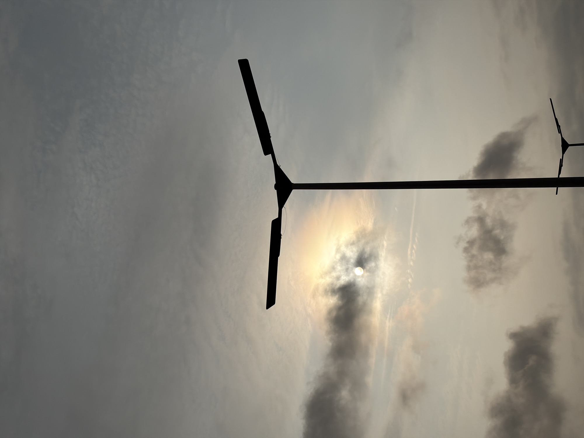 In Alaguntan, Nigeria, a streetlamp is silhouetted against a hazy sky with the sun peeking through clouds. The sun casts a soft, golden glow through the overcast sky, creating a diffused light. The streetlamp has a tall, dark pole with two arms extending outwards, each holding a light fixture, though the fixtures themselves are indistinct silhouettes. The overall scene suggests an outdoor environment in an urban or semi-urban setting, possibly during dawn or dusk due to the quality of light.