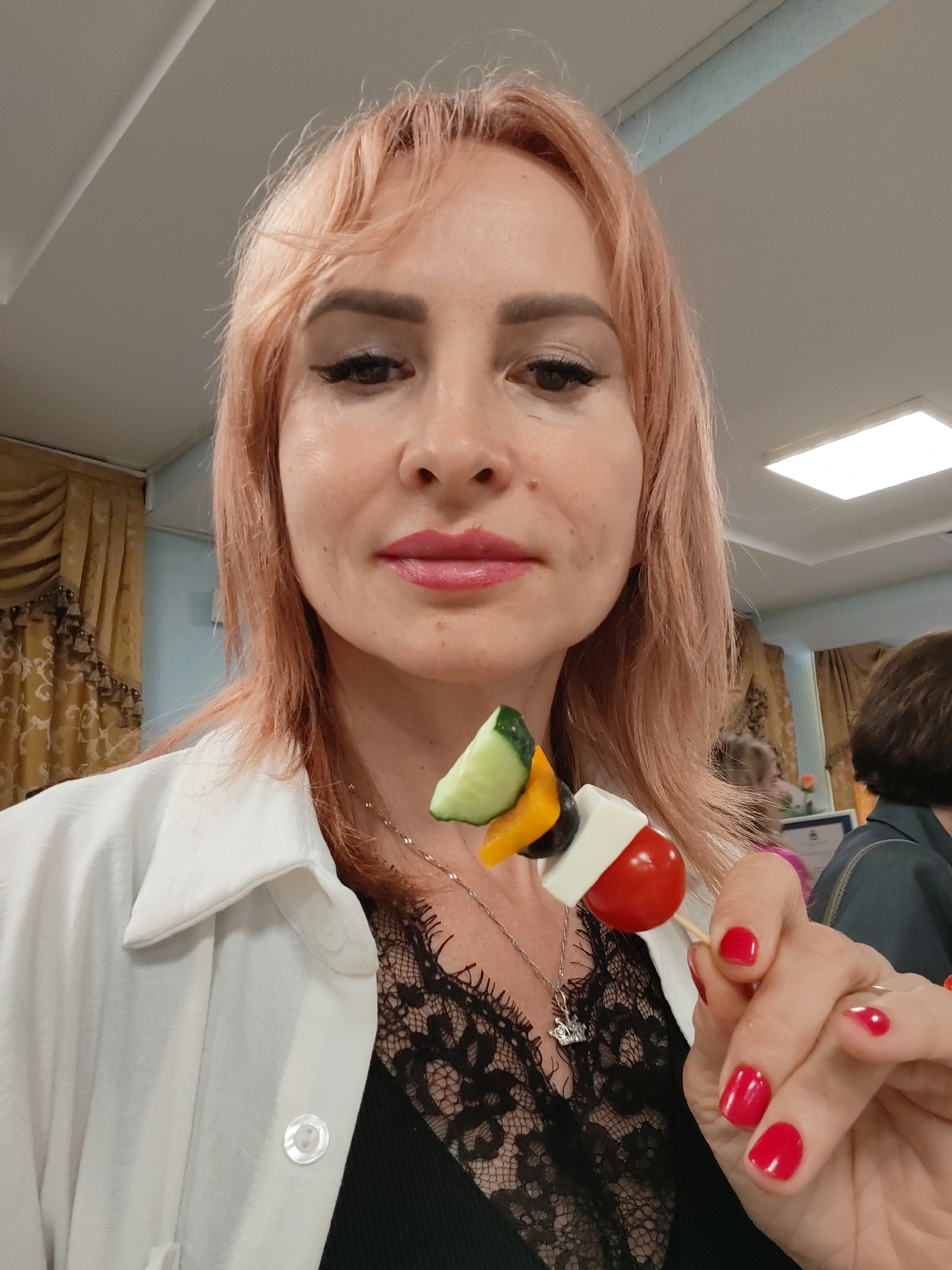 A middle-aged woman with short, pinkish-blonde hair is taking a selfie indoors. She is wearing a white collared shirt over a black lace top, and a silver necklace. She is holding a skewer with cherry tomato, olives, yellow bell pepper, cucumber, and cheese. Her nails are painted red. In the background, there are beige curtains, and other people are visible. The lighting suggests an indoor setting, possibly a community center or a hall in Chaikovsky, Russia, given the details in the background.