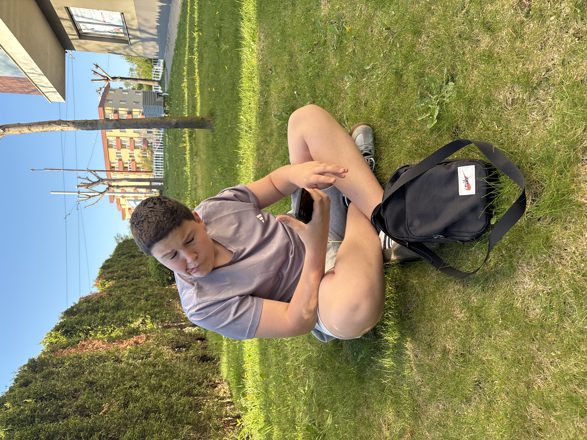 A young person sits cross-legged on a patch of green grass, looking at a smartphone. The person wears a light purple t-shirt, gray shorts, and gray shoes. A black Nike bag rests next to the person on the grass. Behind the person, there are buildings, trees, and a blue sky. Based on the architecture and vegetation, the location could be a park or green space within the city of Sofia, Bulgaria.