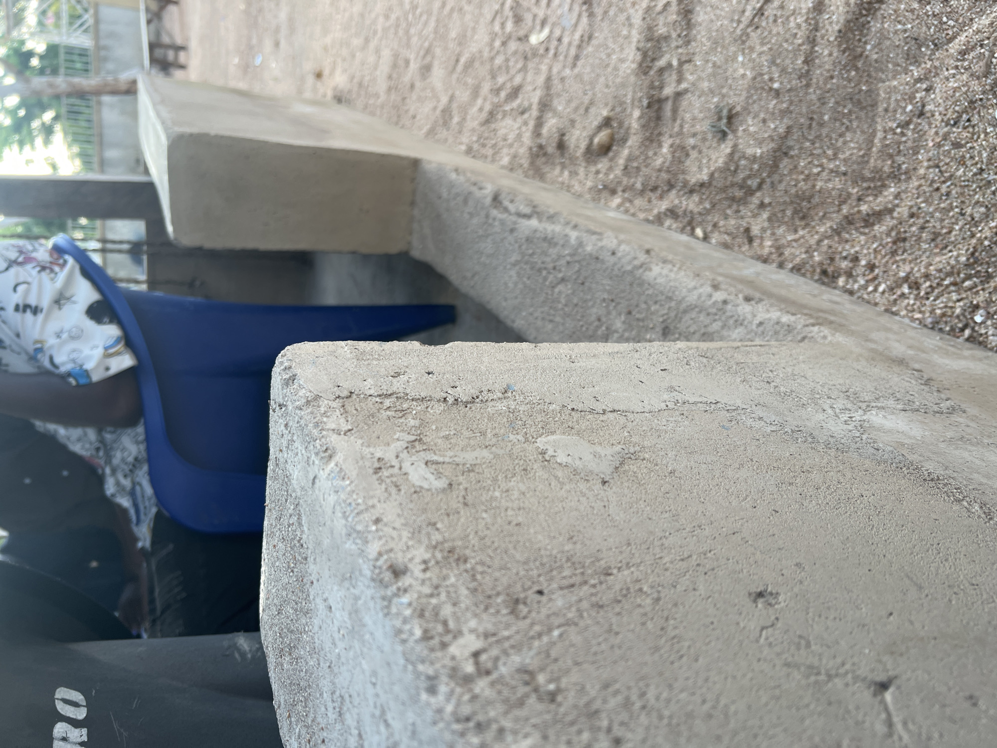 An outdoor, low-angle close-up shot shows a section of a sandy area with two concrete structures in the foreground. The structure on the left is a roughly finished concrete post, angled towards the viewer. To its right is a longer, more smoothly finished concrete wall or ledge. The sandy ground is visible between and beyond these structures, with small pebbles and debris scattered throughout. In the background, partially visible, are two individuals seated on chairs. One person is wearing a blue shirt and the other is wearing a white patterned top. A dark, possibly black object with white lettering is on the far left. Beyond them, a metal fence and trees are visible, suggesting a semi-urban or suburban environment. The lighting indicates it is daytime, with shadows cast on the ground. The scene appears to be in Mayo Dasa, Nigeria, an area characterized by its sandy terrain and common use of concrete structures for everyday purposes.