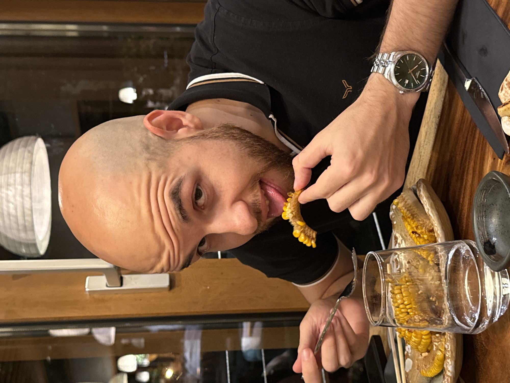 A man with a shaved head, beard, and wearing a black polo shirt with white and brown trim is eating corn on the cob. He is using a fork to hold the corn. A large glass of corn cobs sits on a platter on the table in front of him. The man is wearing a wristwatch on his left wrist. The location appears to be inside a restaurant with wood trim and a view of outdoor lights through the window behind him. It is not possible to identify the city or country from the image.