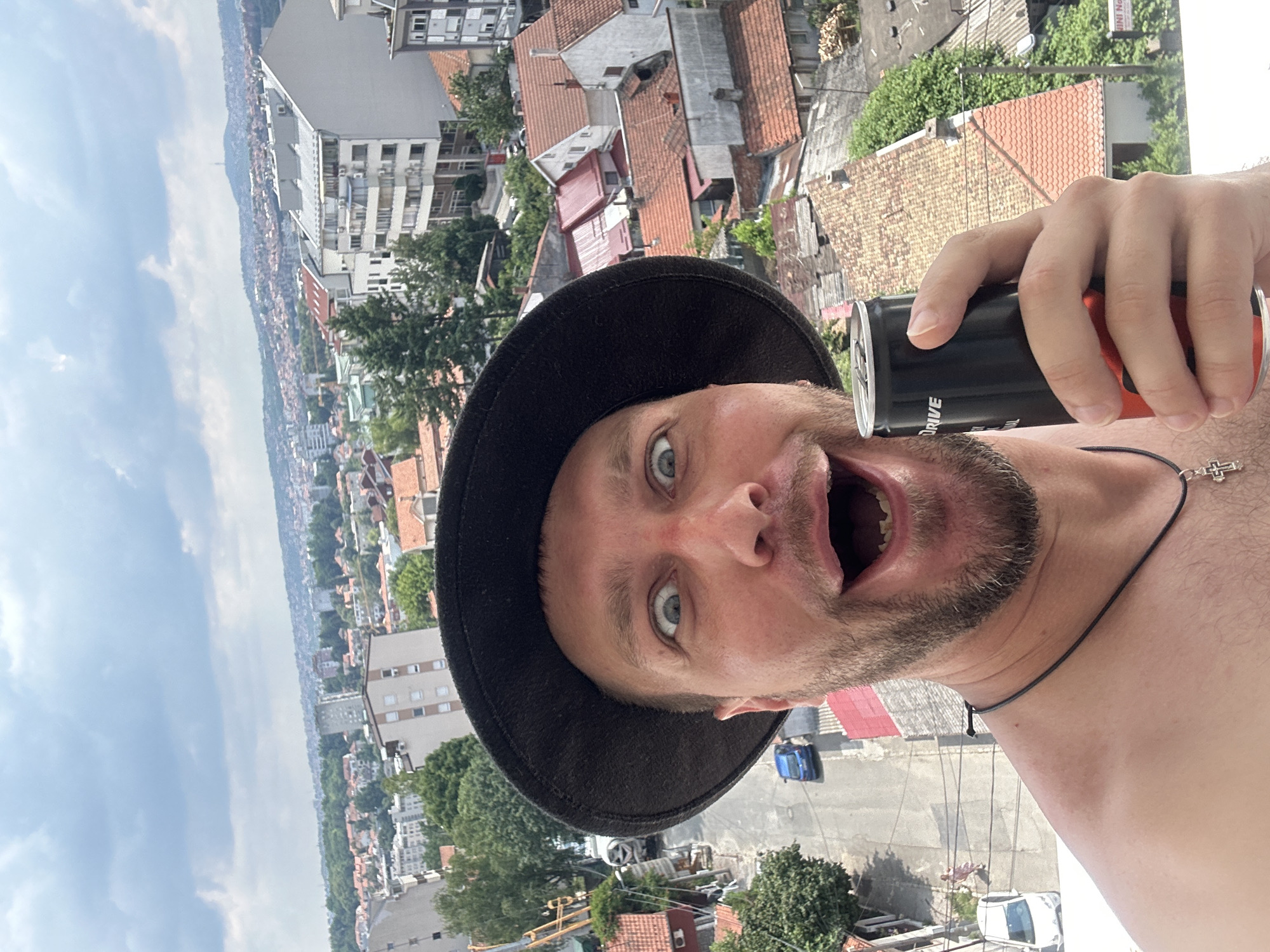 The image is a selfie taken from a high vantage point overlooking a dense urban landscape. A man wearing a black hat and a necklace with a cross is holding a black and red can of energy drink to his mouth. He has a surprised or excited expression on his face, with his mouth wide open and eyes wide. The background shows a sprawling city with numerous buildings, many with red tile roofs, and lush green trees. The sky is partly cloudy, suggesting it could be daytime. The man's arm and shoulder are visible in the foreground, and the can of energy drink has the word "DRIVE" printed on it. The scene appears to be in Zvezdara Urban Municipality, Serbia, as indicated by the provided location context, which suggests a residential or mixed-use urban area. The high angle suggests the photo was taken from a balcony or a rooftop.