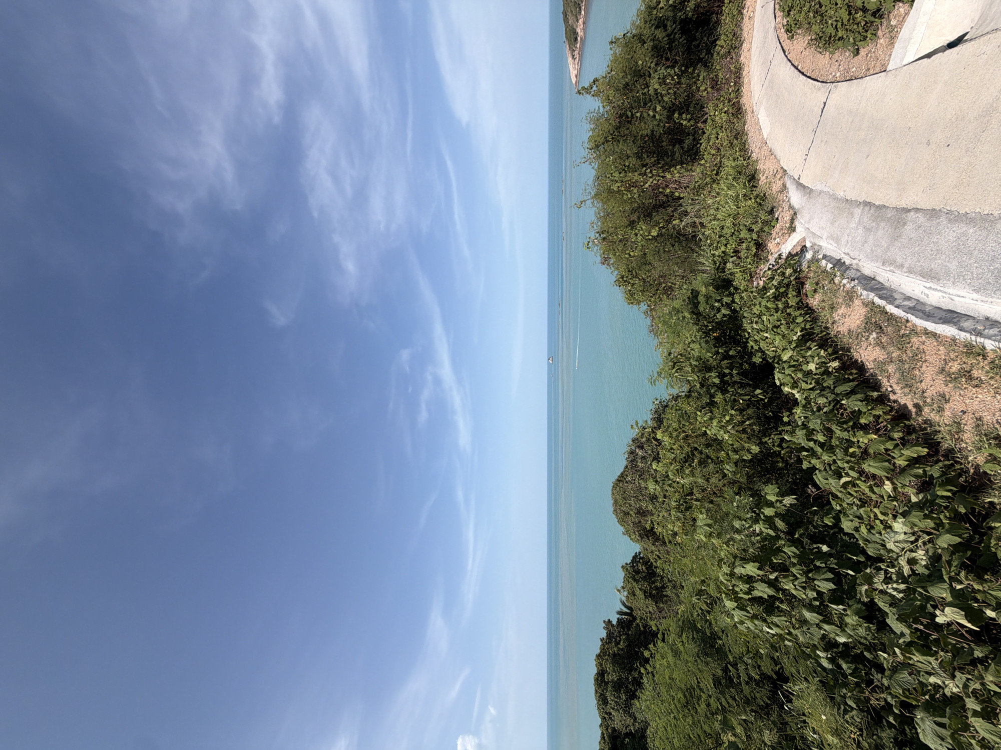 This is an eye-level, medium shot of a coastal pathway on a sunny day in the city of Ko Samui District, Thailand. The pathway, constructed of concrete and gravel, curves to the right, leading the viewer's eye toward the lush, green vegetation that borders it. The vegetation is dense and varied, with trees and bushes of different heights and textures. Beyond the foliage, a vast expanse of turquoise ocean stretches to the horizon, dotted with small boats. The sky above is a clear blue, with a few wispy white clouds adding depth and texture. The overall impression is one of natural beauty and tranquility.