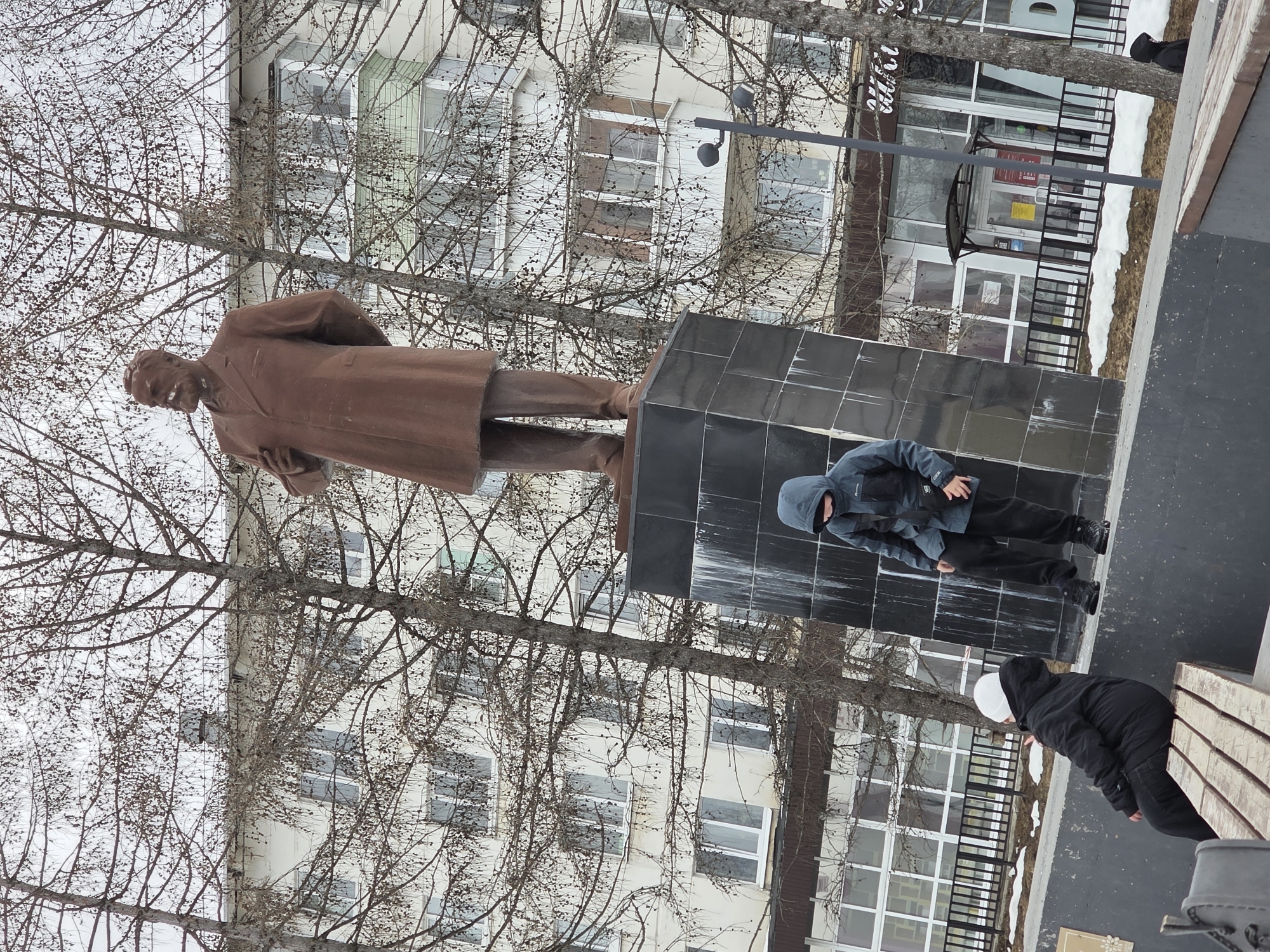 The image depicts the Vladimir Lenin monument in the city of Chaikovsky, Russia. The statue is a full-body bronze depiction of Lenin, standing on a black tiled pedestal. Two individuals are present in the foreground. One, wearing a blue hooded jacket and black pants, is standing near the monument's base. Another individual, dressed in a black puffer jacket and white head covering, is bending near a bench, perhaps tying their shoe. In the background, a multi-story building is visible, with bare trees partially obscuring the view. The scene suggests an outdoor urban setting on an overcast day.