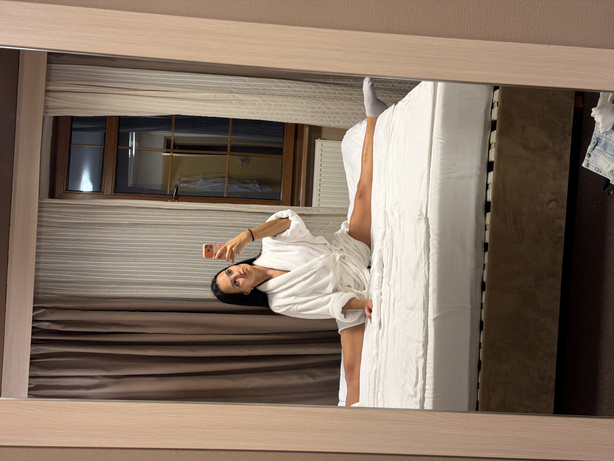 A young woman with dark hair is taking a selfie in a mirror, sitting on a bed in a split position. She is wearing a white bathrobe and grey socks. The room has light-colored curtains and a radiator beneath a window. The bed has white linens. The scene appears to be in a hotel room in Svetlogorsk, Russia.