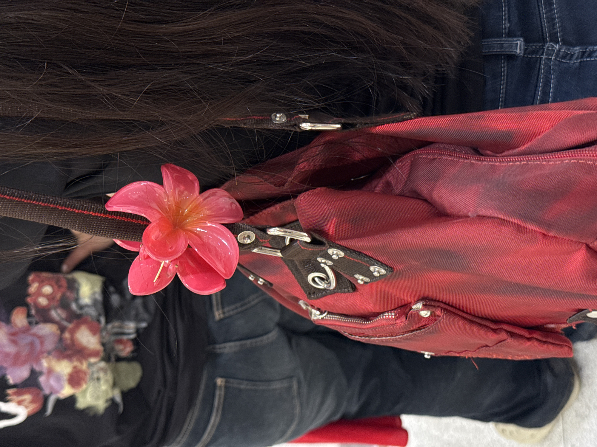 The image shows a close-up of a person's back, from the shoulders down, with a red bag hanging from their shoulder. The person is wearing a black t-shirt with a floral print and blue jeans. A bright pink flower accessory is attached to the strap of the bag. The background is blurred, but it appears to be an indoor setting, possibly a shopping mall or a public building. There is no visible indication of the city of Tbilisi, Georgia.