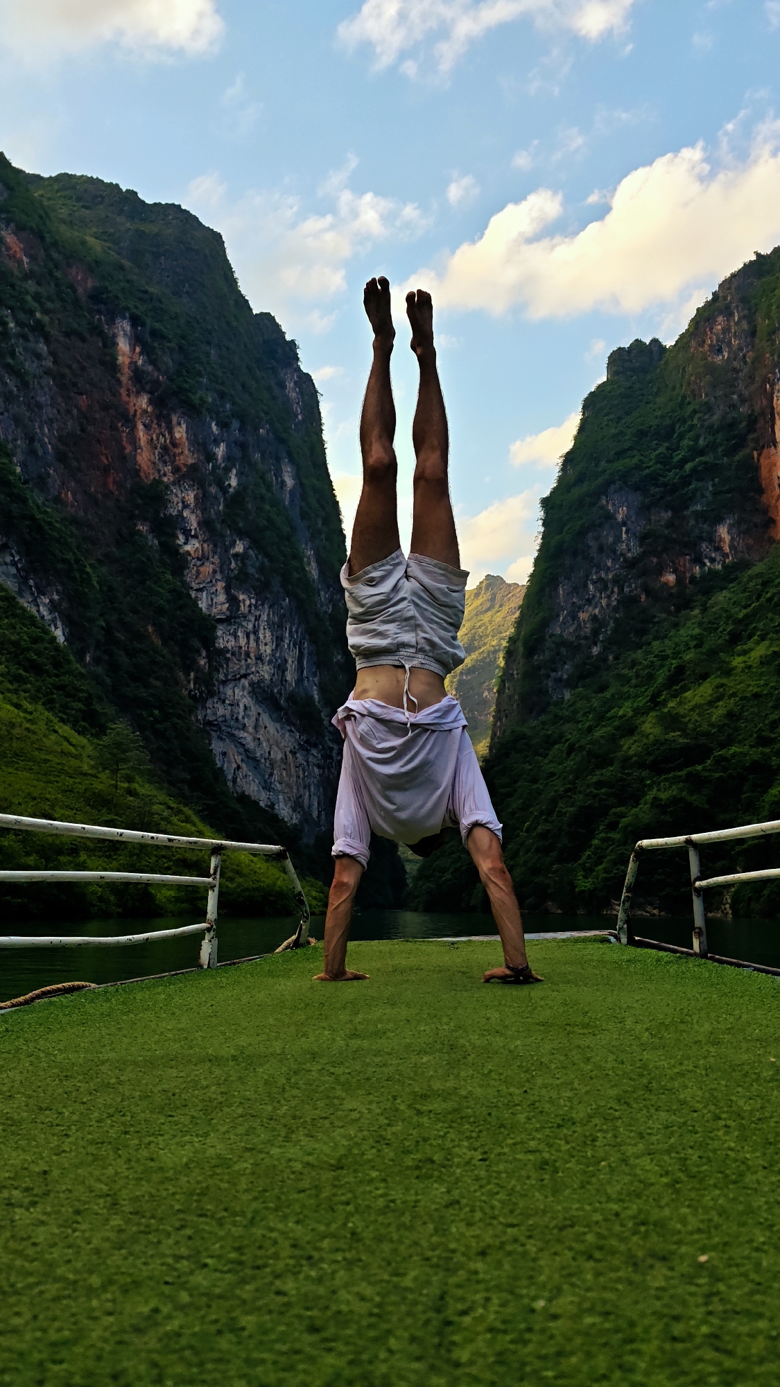 A man is performing a handstand on a green platform with white railings, likely a boat deck, in a narrow river gorge. Tall, steep, heavily forested mountains rise on either side, with rocky cliffs visible. The sky is partly cloudy, with blue patches. The man is wearing light-colored shorts and a t-shirt that has ridden up during the handstand. The photo appears to have been taken in Đồng Văn Commune, Vietnam, given the landscape's characteristics.