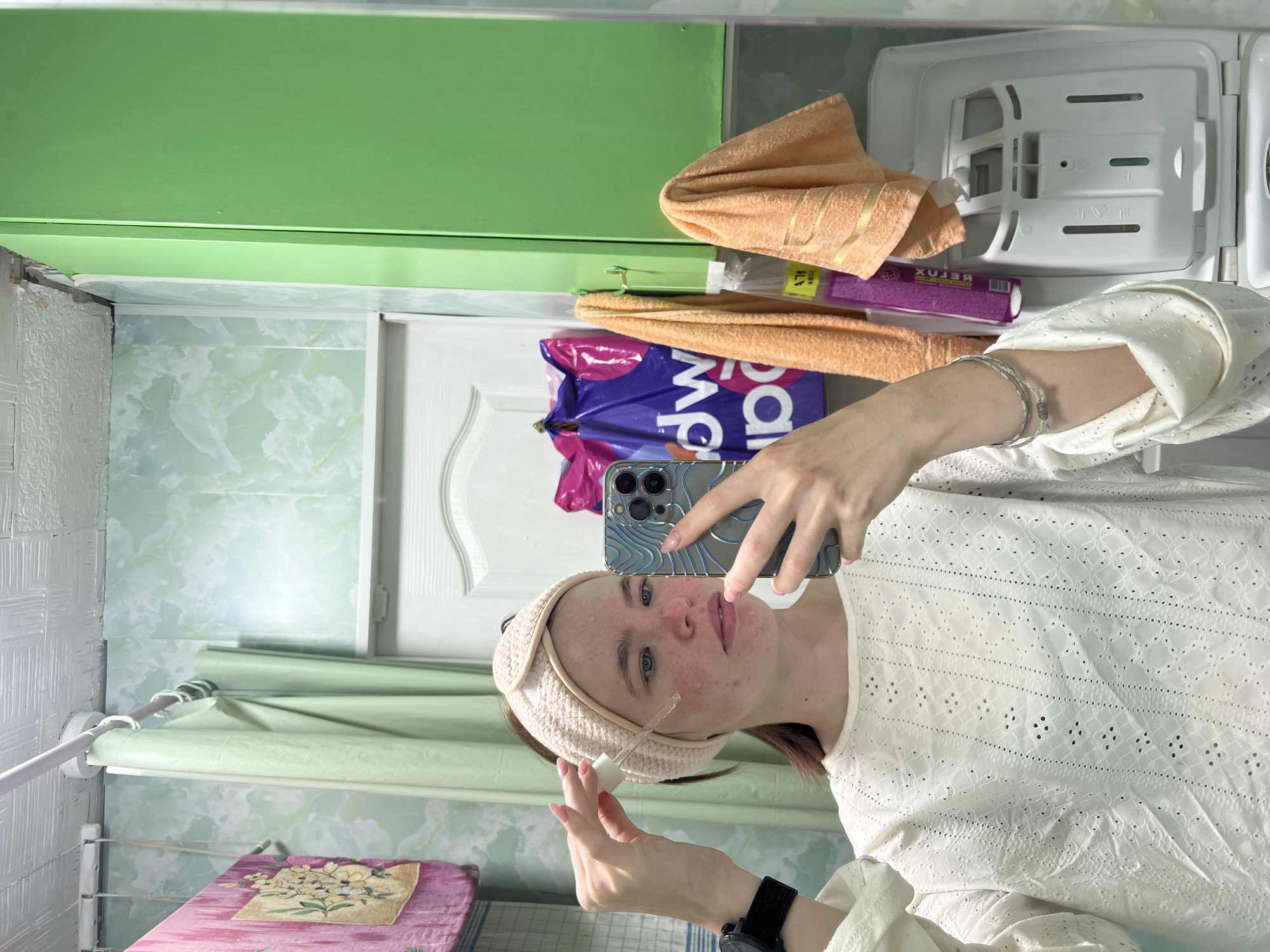 A young woman is depicted taking a mirror selfie indoors, likely in a bathroom or utility room in Chaikovsky, Russia. She wears a light-colored, possibly white or cream, long-sleeved top with eyelet embroidery and a light pink or beige headband around her head. Her face is visible, showing some skin blemishes. She holds a smartphone with a textured case in her left hand, using it to take the picture, and a dropper bottle, possibly containing a skincare serum, in her right hand, suggesting she is in the midst of a beauty or self-care routine. She has a watch on her left wrist and bracelets on her right.
The background, seen through the mirror's reflection, reveals a domestic setting. Visible elements include light green walls with a marbled pattern, a white door with decorative molding, and a green shelf or cabinet. Household items include an orange towel, a purple plastic shopping bag with "Ma" visible on it (possibly from a store like "Magnet"), and a white storage unit or refrigerator compartment. A purple product package with the reflected text "BETOX" (likely "DETOX") is partially visible behind the towel. Further back, a white drying rack holds a pink patterned fabric, possibly an ironing board cover, next to a light green curtain or blind. The scene captures a private moment in a home environment.
