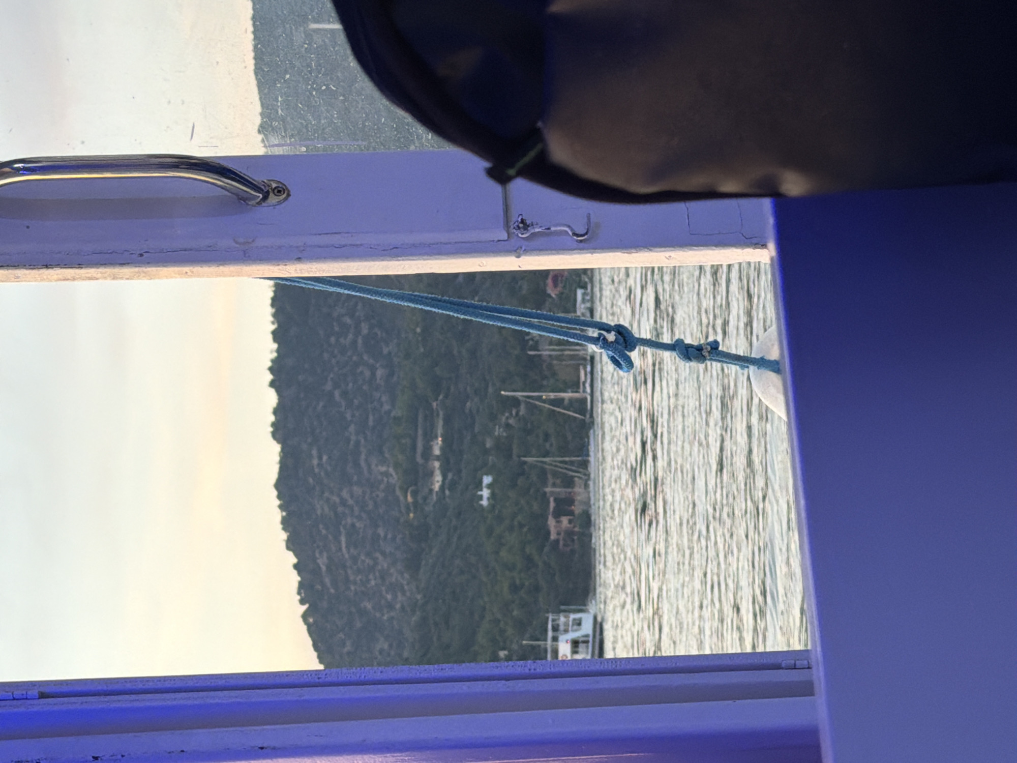 The image, when correctly oriented, presents a scenic view from inside a boat or structure looking out onto a body of water and a distant landmass.
In the foreground, a white horizontal frame, likely part of a window or boat railing, spans the width of the image. On the left side of this frame, a shiny, chrome-plated grab handle is mounted. Towards the right, a small, intricate metal hook-latch is visible. Below the frame, a blue braided rope, tied in a knot, extends downwards into the water. Above the frame, a dark blue or black object, possibly a boat cover or a bag, partially obscures the top left corner. On the far right and bottom, elements of a vibrant blue/purple color suggest interior lighting or painted surfaces of the boat.
The midground features a calm body of water with subtle ripples and reflections, indicating a serene environment. In the distance, several boat masts are discernible, suggesting a harbor or anchorage.
The background is dominated by a large, verdant mountain or island, covered densely with trees and vegetation. Small, light-colored buildings are scattered across the mountainside. Above the mountain, the sky is a soft, pale yellow and white, indicative of either early morning or late afternoon light.
The scene depicts a peaceful moment, possibly during sunrise or sunset, while onboard a vessel. The overall impression is one of tranquility and natural beauty, consistent with the coastal landscape of Poros, Greece. No discernible text is visible in the image.