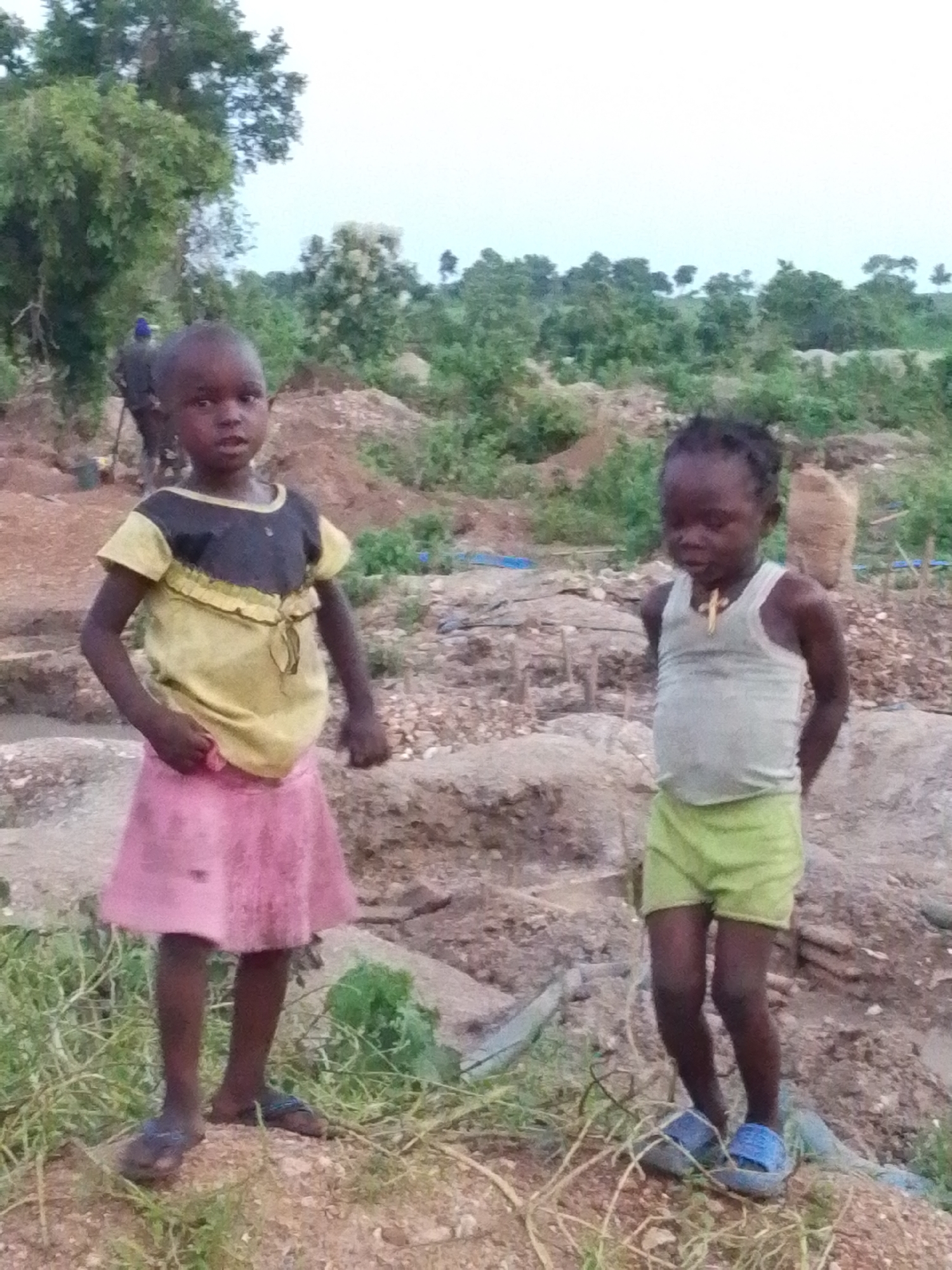 Two young children are standing in an outdoor, arid environment with sparse vegetation and eroded earth. The child on the left, a girl, is wearing a yellow and black t-shirt with a pink skirt and blue sandals. She is looking towards the camera with her hands by her side. The child on the right, also a girl, is wearing a light grey tank top with light green shorts and blue sandals. She has a necklace with a gold cross pendant and is looking down with her hands behind her back. In the background, there are trees and a hazy sky. The ground is covered in loose soil and rocks, suggesting a construction or mining site. The location is identified as Otulu, Nigeria.