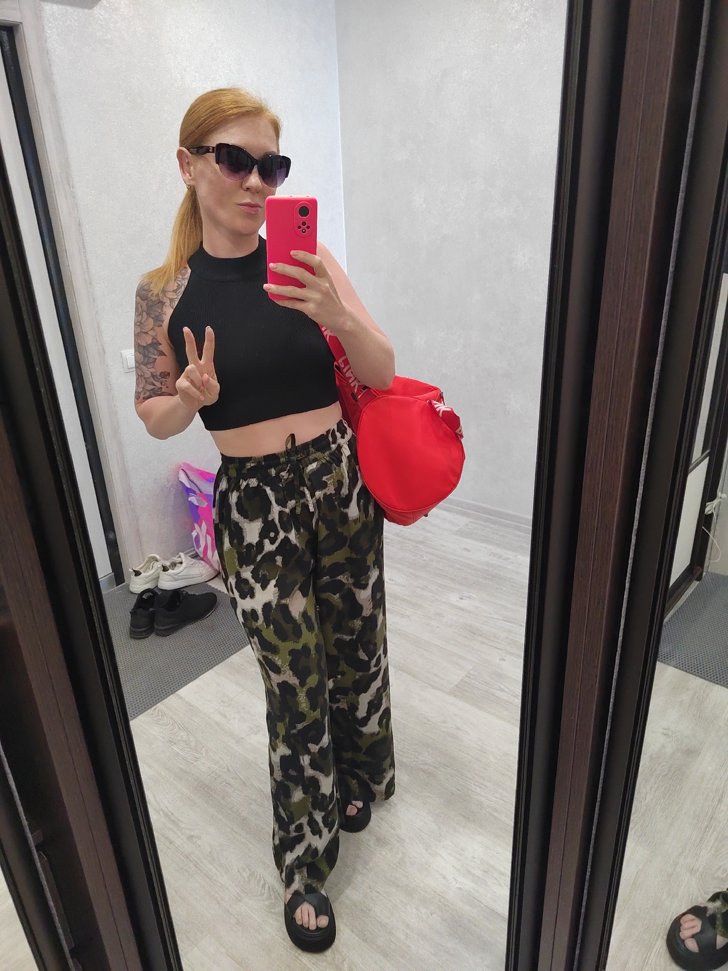 A woman with red hair in a ponytail, wearing sunglasses, a black crop top, and patterned pants, stands in a room and takes a selfie in a mirror. She is holding a pink phone in her right hand and making a peace sign with her left hand. A red bag is slung over her left shoulder. In the reflection of the mirror, white sneakers and black shoes are visible on the floor. The room appears to be a dressing area or a room with multiple mirrors. The floor is light-colored wood. The walls are white. The overall impression is of a casual setting, possibly a home or a fitting room. The city of Saint Petersburg, Russia is not discernible from the image.