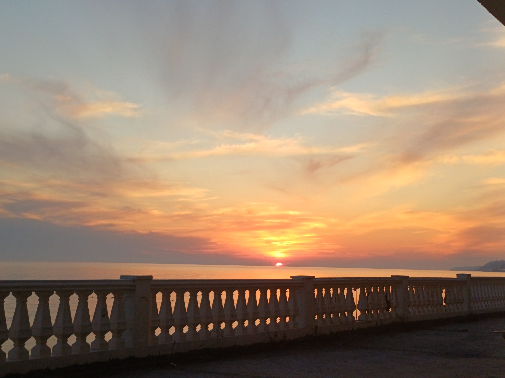 The image displays a sunset over the Black Sea, viewed from a coastal promenade in Sochi, Russia. The sun, a prominent orange-red semi-circle, is positioned on the horizon line, partially submerged below the calm sea surface. The sky above is a gradient of colors, transitioning from a light blue-grey at the top to vibrant oranges, yellows, and reds closer to the horizon, with cirrus and altocumulus clouds illuminated by the setting sun. The sea reflects the warm hues of the sky, appearing mostly calm with minimal wave activity.
In the lower half of the frame, a long, light-colored balustrade stretches horizontally. This balustrade, likely constructed from concrete or stone, features a series of decorative balusters with distinct inverted teardrop or diamond-shaped openings, supporting a flat top rail. It is presented in silhouette against the brighter sunset, casting it in shadow. The ground directly in front of the balustrade is a dark, rough-textured surface, possibly paved or gravel. A distant landmass is faintly visible on the far right horizon. No people or active subjects are present in the scene.