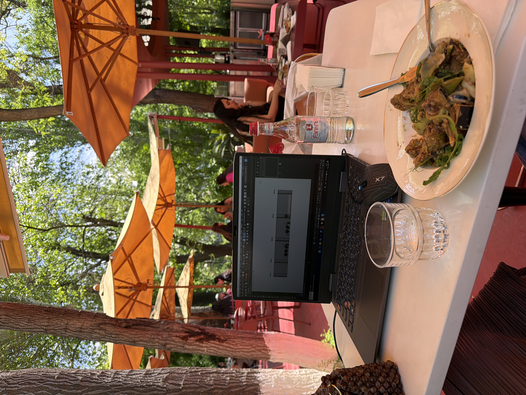 Captured outdoors in Yerevan, Armenia, the image presents a white tabletop dominated by a laptop displaying a CAD program interface. Beside the laptop is a clear glass tumbler, and an open plate of salad is placed on the opposite edge. A small glass bottle of Armenian "BYUREG" mineral water sits beside the laptop. A few stacked white napkins are in the frame. The background is filled with tables and chairs, along with other people. Multiple orange parasols provide shade. Tree trunks in the foreground add depth to the composition.