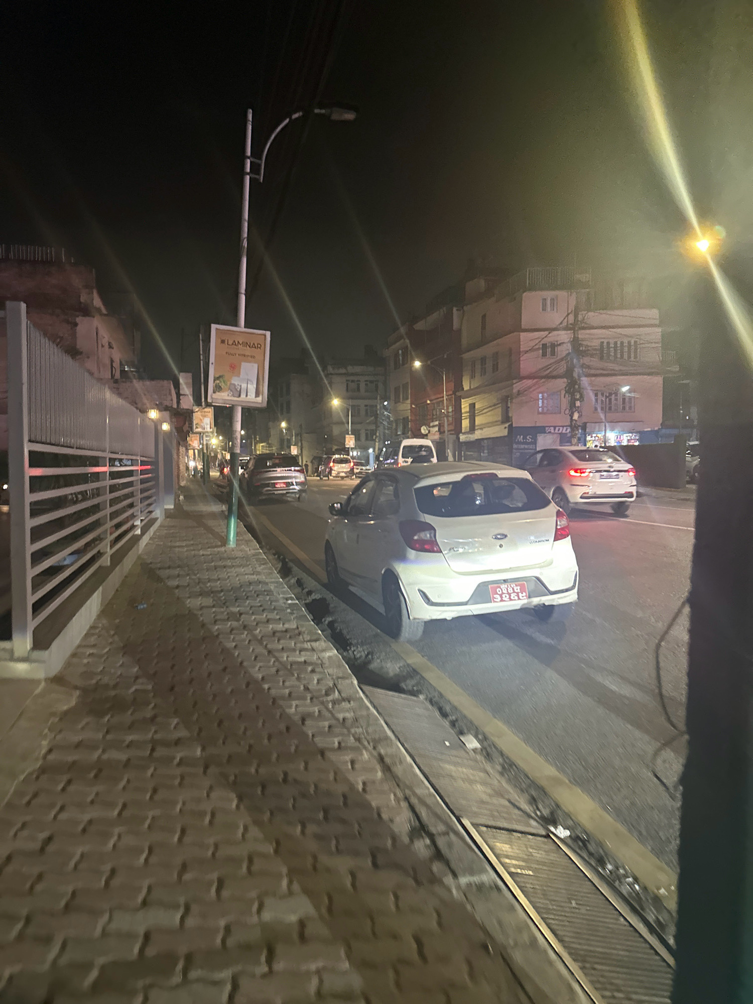 The image shows a street scene at night in Kathmandu Metropolitan City, Nepal. A white Ford Figo is parked on the right side of the road, with its rear facing the camera. The car has a red license plate with Nepali script. Another white car, a Hyundai i10, is visible further down the road, also parked on the right. A street light illuminates the scene from above. The sidewalk on the left is paved with interlocking bricks. A lamppost with a sign that reads "LAMINAR FULLY VITRIFIED" is also visible. Buildings line the street on both sides. The overall impression is of a busy urban street at night.