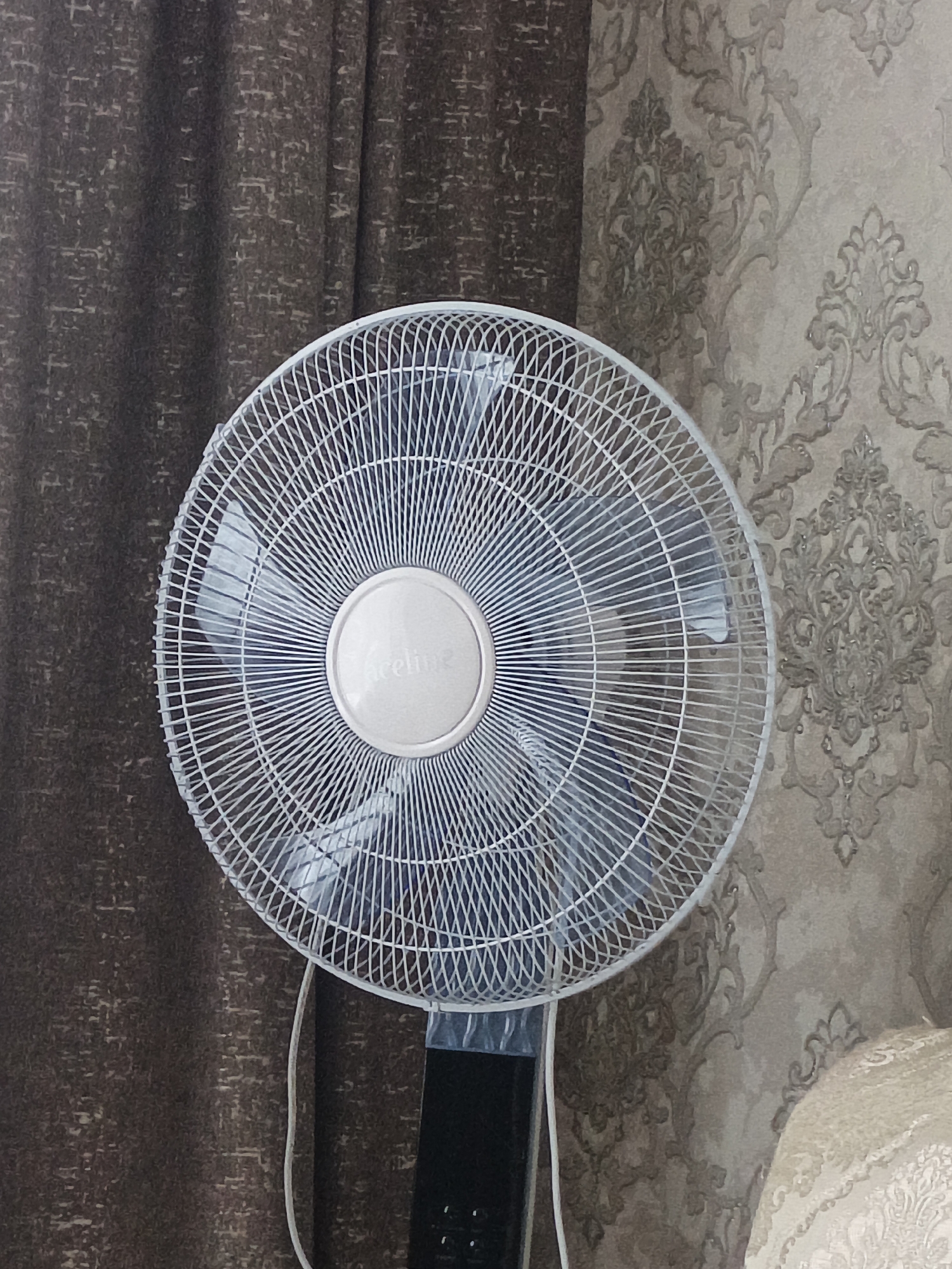 A white oscillating fan is positioned in the foreground, occupying the majority of the frame. The fan has a silver circular center with faint text on it. Behind the fan, on the left, is a dark brown curtain with a textured pattern. To the right of the fan, the background consists of a patterned wallpaper in beige and taupe tones, featuring ornate floral motifs. The lighting appears to be indoor and diffused, casting soft shadows. The image does not provide any information that would allow for its location to be identified as Sukhodol, Russia, or any other specific city.