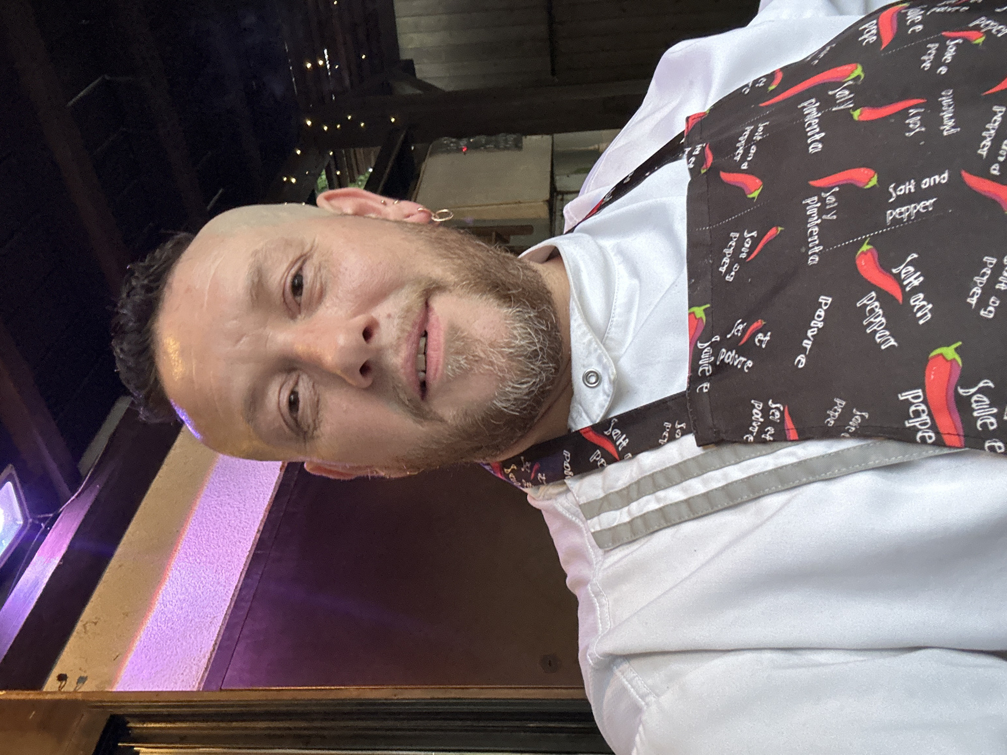 A man with short hair and a beard is wearing a white chef's jacket and a black apron with red chili peppers and the words "salt and pepper" printed on it. The man appears to be standing outdoors at night, based on the ambient lighting and visible elements of the surroundings, which may be Новоглаголево, Russia.