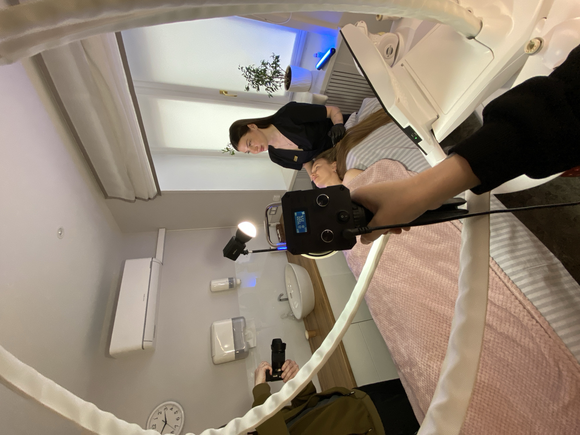 This image captures an indoor scene, likely within a beauty salon or aesthetic clinic in Votkinsk, Russia. The setting is a clean, well-lit treatment room with light-colored walls.
The primary activity depicted is a beauty or aesthetic treatment being performed on a client. A woman, dressed in a black uniform and black gloves, leans over another woman who is lying relaxed on a treatment bed covered with pink and striped linens. The practitioner appears focused on the client's face or head area, suggesting a detailed procedure.
Simultaneously, the entire process is being professionally photographed or videographed. In the foreground, a third person, wearing a dark green jacket, holds a professional camera with a large lens, aiming it at the two women. Adjacent to the camera, a piece of lighting equipment, possibly an LED panel or ring light, is visible, illuminating the scene. Its digital display shows "CCT 5500K 35%", indicating a color temperature of 5500 Kelvin and 35% brightness, suggesting a controlled lighting setup for media capture. Another smaller light source is positioned on a stand, casting light near the sink area.
Notable objects in the room include a white sink with a tap set into a wooden counter, wall-mounted dispensers (likely for soap or sanitizer), and a white air conditioning unit mounted high on the wall. A window with white blinds is visible, on its sill sits a potted plant and a blue light fixture. A wall clock, partially visible, indicates a time around 12:30 or 1:30. The shot itself is framed by white, flexible tubes, possibly belonging to treatment equipment or serving as aesthetic elements. The overall atmosphere suggests a professional environment focused on client care and content creation.