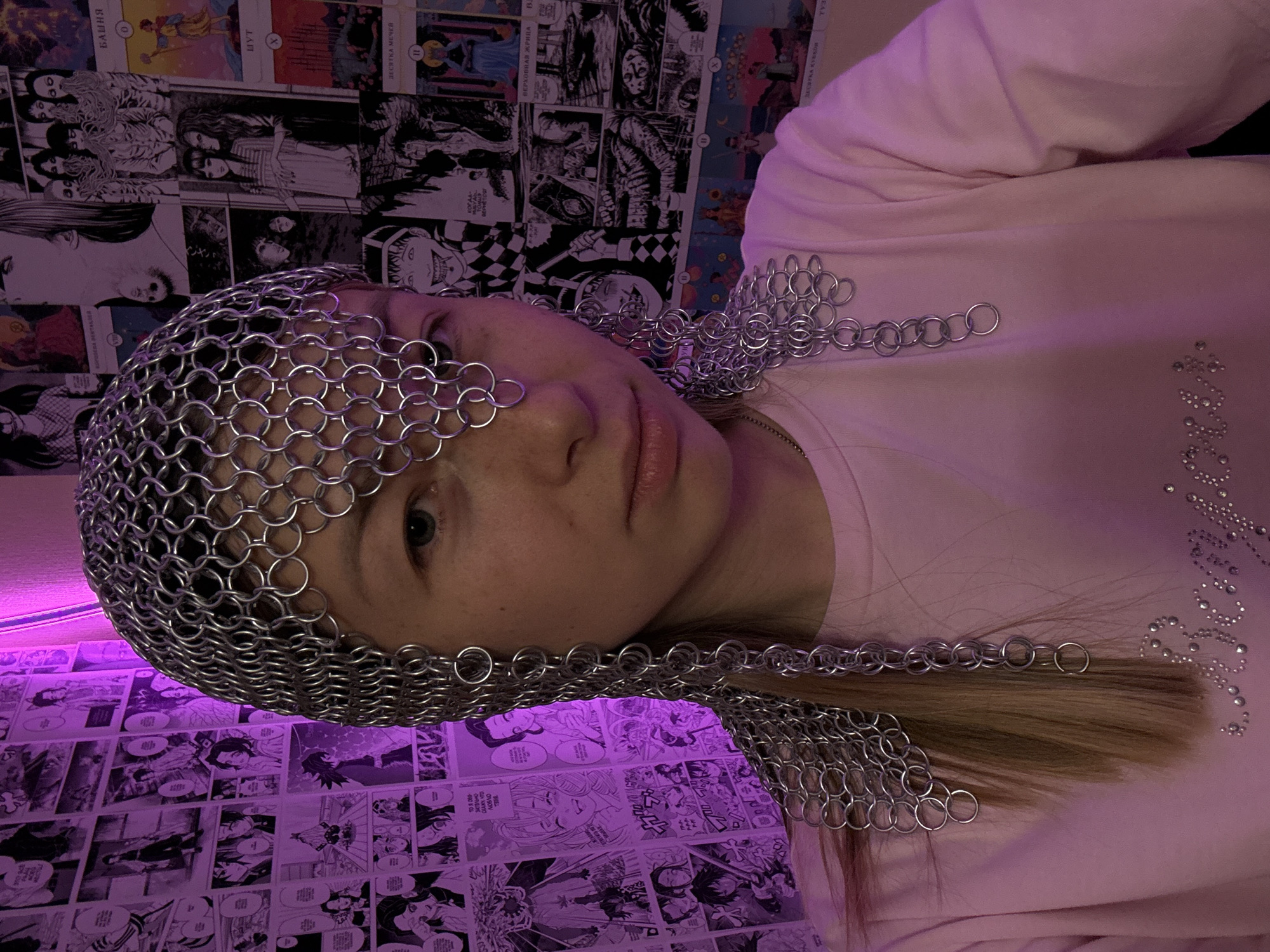 The image captures a young woman in a medium shot, presenting a self-portrait within an indoor setting. She has light-toned skin, light blue-green eyes, and light brown hair styled to fall over her shoulders. Her expression is neutral, and she gazes directly into the camera. She wears a light pink long-sleeved top adorned with the word "Baby" spelled out in rhinestones on the lower front. A thin, silver-colored chain necklace is partially visible around her neck.
A unique headpiece, resembling chainmail and constructed from numerous interconnected silver-colored metal rings, covers the top of her head, forehead, and extends in decorative strands down the sides of her face to her shoulders. A small section of this headpiece rests over the bridge of her nose.
The background features a wall densely covered with printed pages. These pages primarily consist of manga panels, identifiable by their distinctive art style, including elements reminiscent of the "Demon Slayer" series. Additionally, several prints depict Tarot card illustrations, with visible Cyrillic text identifying cards such as "БАШНЯ" (Tower), "ШУТ" (Fool), "ВЕРХОВНАЯ ЖРИЦА" (High Priestess), and "ДЕСЯТКА МЕЧЕЙ" (Ten of Swords). Further Cyrillic text is present within manga speech bubbles, confirming a Russian-speaking cultural context for the environment. The background is bathed in a prominent purple ambient light, casting a magenta hue across the decorated wall. The scene is located in the city of Moscow, Russia.