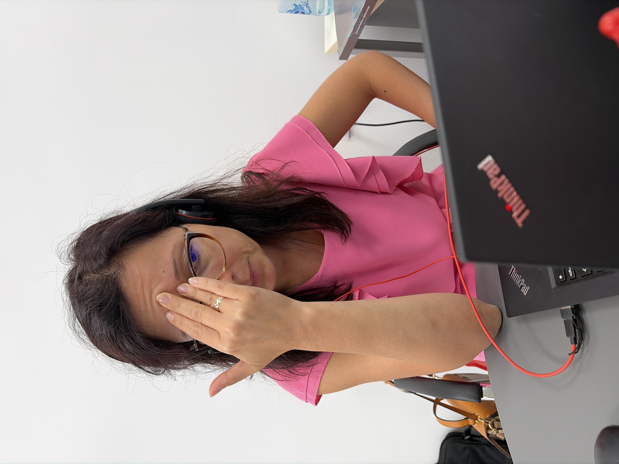 A woman with dark hair and glasses is sitting at a gray desk, wearing a pink shirt and headphones. She is holding her hand to her forehead, covering her eye. A black laptop with a red "ThinkPad" logo is in front of her, connected to a red cable. A brown purse is on the left side of the desk. The background is a white wall.