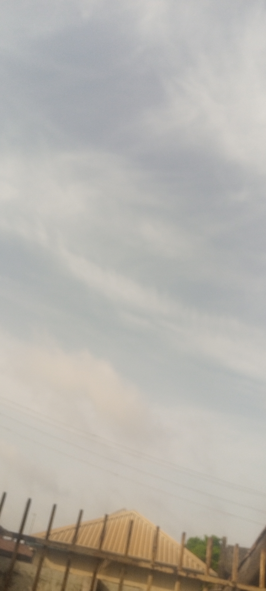 The image shows a cloudy sky above buildings in Sabon Tasha, Nigeria. There are several buildings with corrugated metal roofs under construction and electrical wires are visible against the sky.