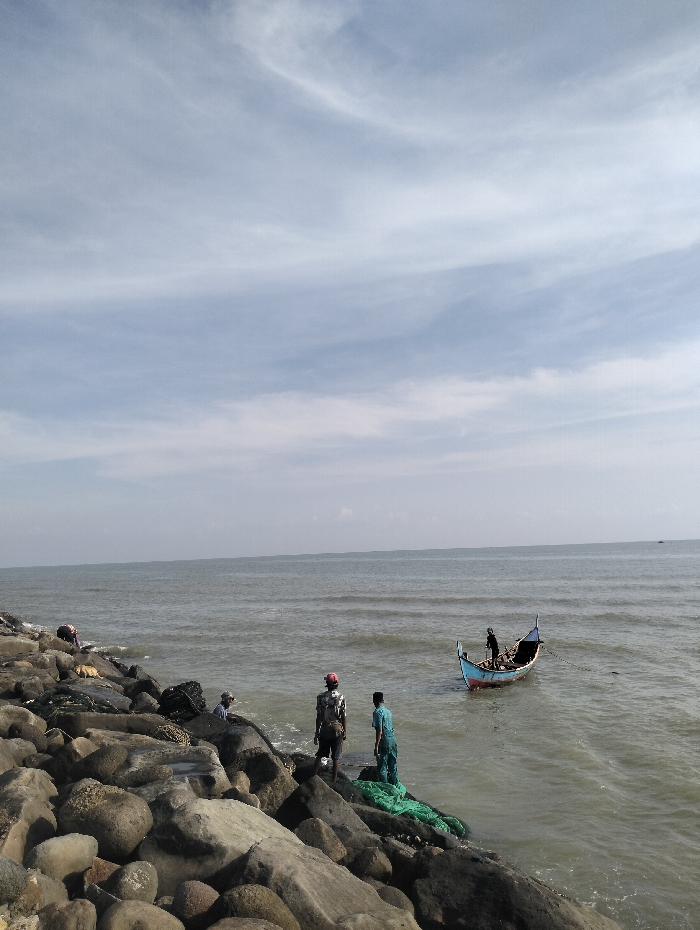 On a rocky shore, several people are near the water's edge. Some stand on the rocks, near green netting, while others are further down towards the sea. A small, colorful boat is anchored in the water, with figures aboard. The sea extends towards the horizon under a bright, slightly cloudy sky.