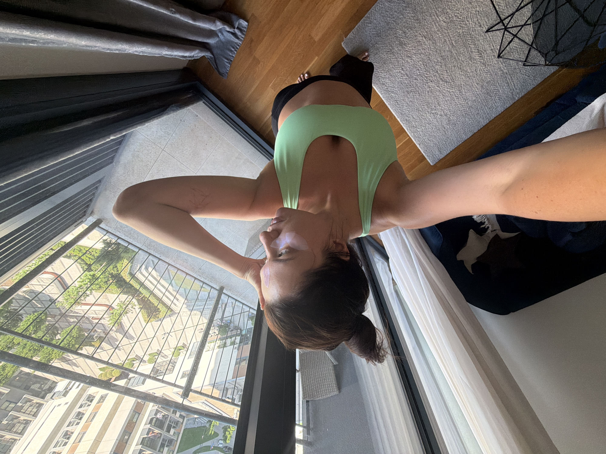 In Belgrade, Serbia, a woman with dark hair pulled into a bun and wearing eye patches stands on a wooden floor inside an apartment. She is dressed in a light green sports bra and black leggings, with her left arm raised to her forehead. The room opens to a balcony overlooking an urban courtyard with trees and modern buildings. Sunlight streams in, casting shadows and highlighting the scene.