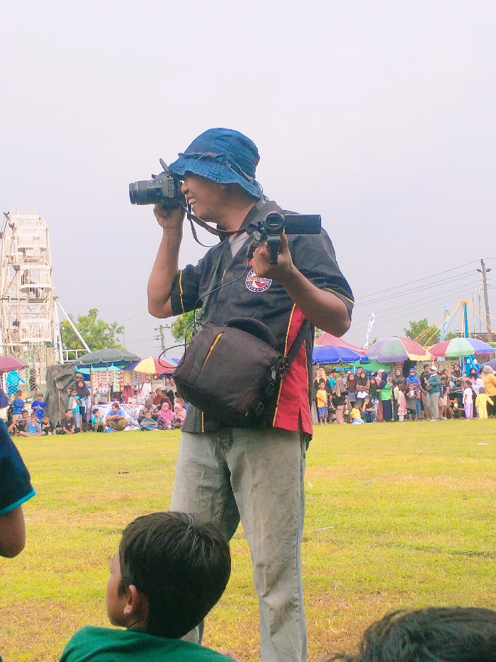 A man wearing a blue hat and a red and black shirt is holding a camera and a camcorder. He is standing in a field of green grass, looking at something to the right of the frame. Behind him, there is a large crowd of people, many of them women wearing hijabs, gathered on the other side of the field. In the background, a large white Ferris wheel can be seen partially obscured by trees. There are colorful tents and canopies to the left of the man, behind him. The man's camera is pointed toward the crowd of people. The image was taken in Karangmalang, Indonesia at coordinates -7.47 latitude and 111.064 longitude.