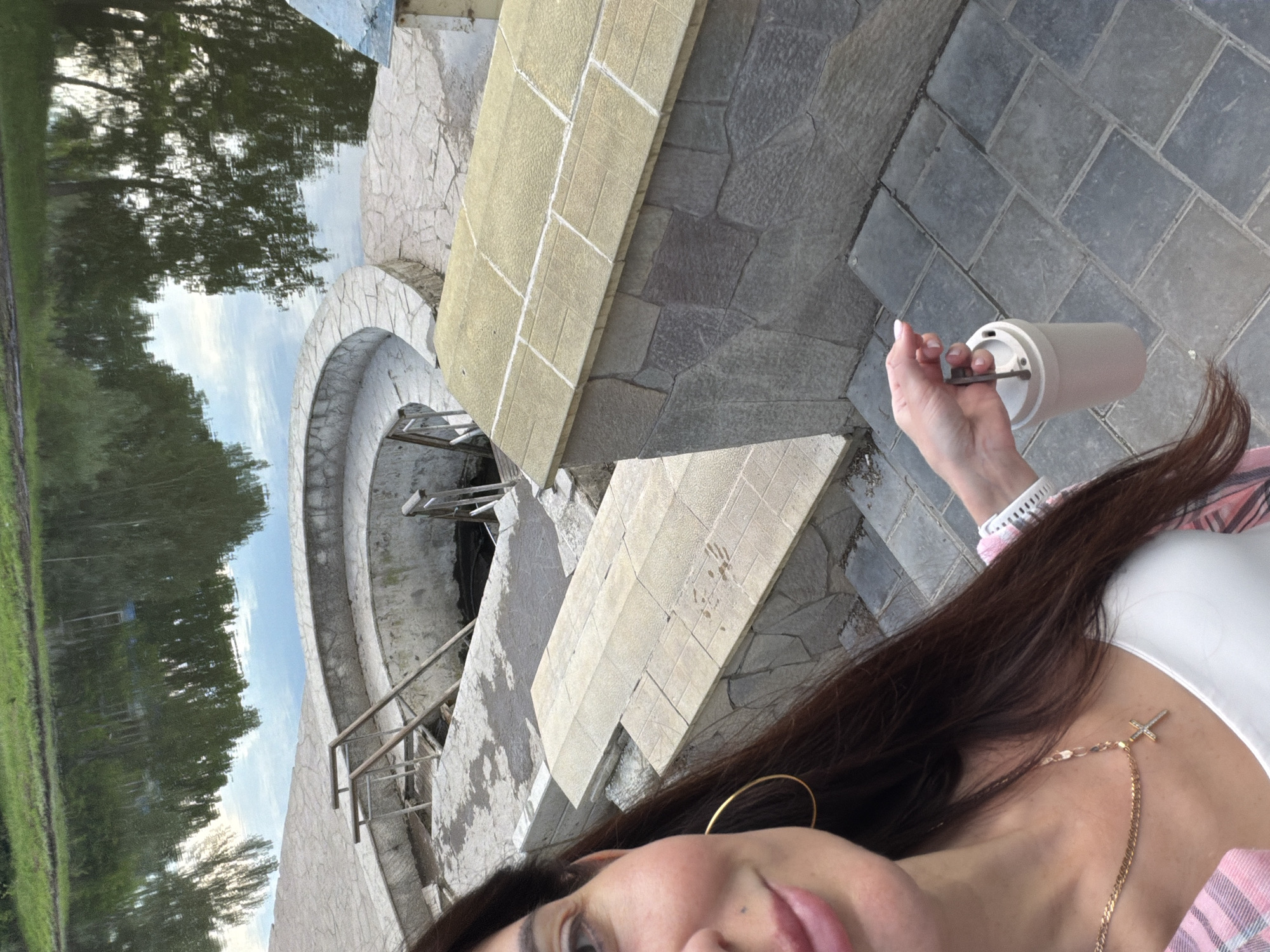 A woman, with long dark hair and wearing a pink and white plaid shirt and a white top, stands in front of a body of water, holding a travel mug. She is wearing large gold hoop earrings and a gold necklace with a cross pendant. Behind her, a circular structure made of stone and concrete surrounds a dark opening that leads into the water. Steps made of stone slabs lead down to this structure. The water, reflecting trees and sky, is calm and appears to be a natural body of water. The overall scene suggests an outdoor location, possibly a park or a public bathing area, in Volzhskiy, Russia.
