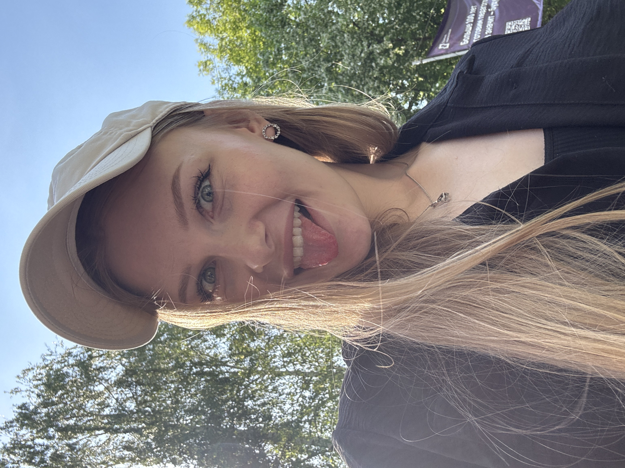 The image is a selfie of a young woman outdoors, with a partially visible green tree canopy and a section of blue sky in the background. The woman is wearing a light-colored cap and a dark top, and a silver necklace. She is sticking her tongue out, smiling, and appears to be in the city of Perm, Russia.