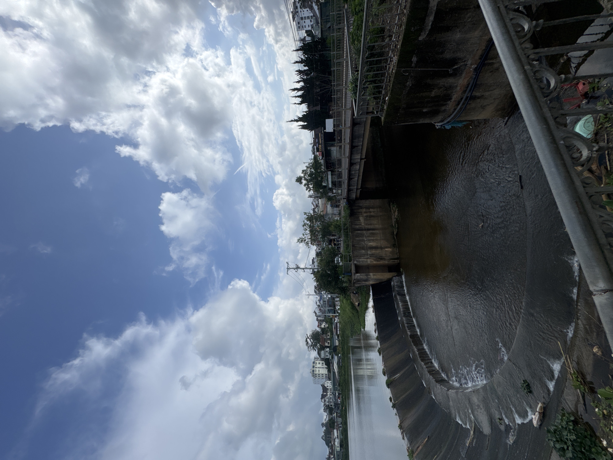 This daytime image, taken in Lâm Viên - Đà Lạt, Vietnam, captures a river or canal flowing through an urban environment under a partly cloudy sky. The sky features distinct white cumulus clouds against patches of clear blue.
In the foreground, the dark water of the river occupies the lower portion of the frame. A concrete weir or small dam is prominent, with water cascading over its edge, forming white foamy patterns. On the immediate right, a grey metal railing, possibly belonging to a bridge or viewing platform, is visible.
A concrete bridge spans the water further along the river, connecting the two banks. Both sides of the river are lined with a mix of buildings, including multi-story structures and smaller houses, interspersed with numerous green trees and bushes. Power lines and utility poles can be seen above the buildings on the left bank. Underneath the bridge on the right bank, some indistinct objects, including what appears to be a light blue/green plastic item and reddish material, are visible near the water's edge.
No people or specific activities are depicted, and there is no discernible text. The scene presents a calm, everyday view of a developed waterway in a town or city.