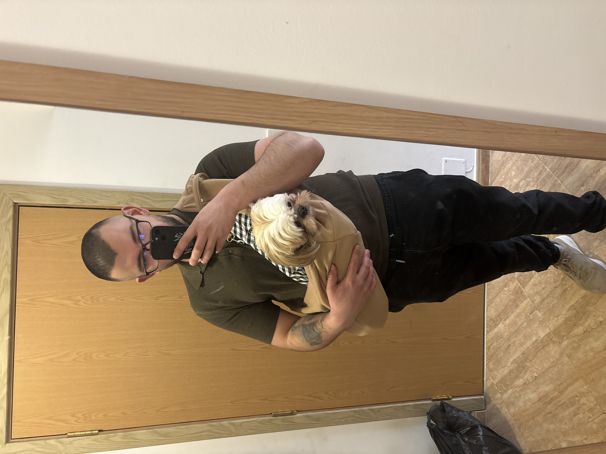 A man is taking a mirror selfie while holding a small, long-haired dog in a beige carrier against his chest. The man is wearing an olive green short-sleeved polo shirt and black jeans. He has dark hair, a beard, and glasses. He is holding a black smartphone with a black case to take the picture. His left arm has a visible tattoo. The floor is covered with light brown tiles. In the lower right corner, there is a black garbage bag. The mirror reflects a wooden door and white wall. The location cannot be determined from this image.