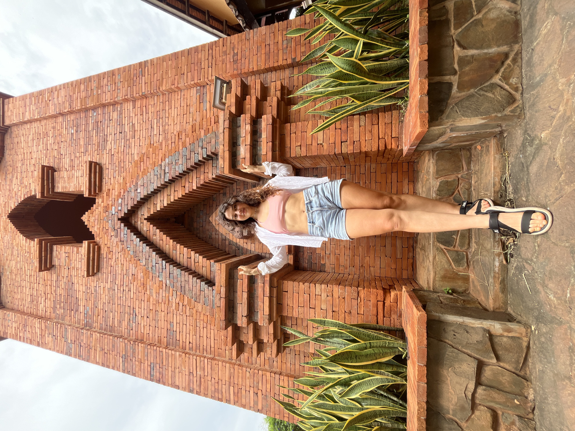 A young woman stands in front of a tall, ornately decorated brick structure with a stepped, pointed archway. She is wearing a pink crop top, denim shorts, a white open shirt, and black sandals. Snake plants are visible on either side of the entrance. The woman is smiling and has her hands raised slightly. The architectural style suggests it could be a temple or a historical building in Phu Thuy Ward, Vietnam. The sky is overcast.