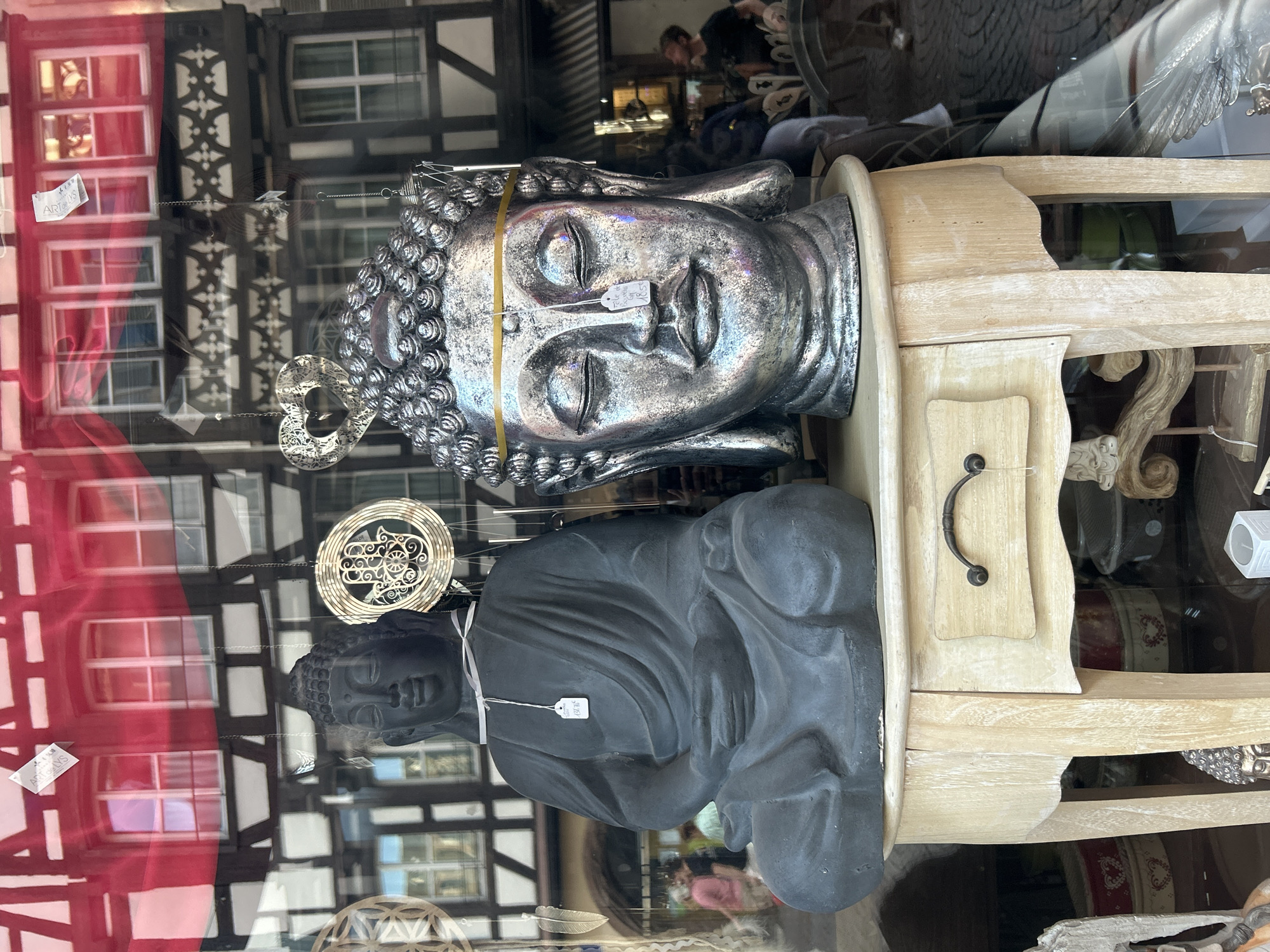 A shop window display in Strasbourg, France, features two Buddha statues – one silver and one black – positioned next to a light-colored wooden side table. The statues and table are viewed through the glass of the shop window, which also reflects the facades of buildings across the street. Details such as window frames and decorative elements on the reflected buildings are visible. The shop's interior and figures within it are also faintly visible through the reflections. The presence of price tags on the statues suggests they are for sale. Other decorative items are visible within the store's display.