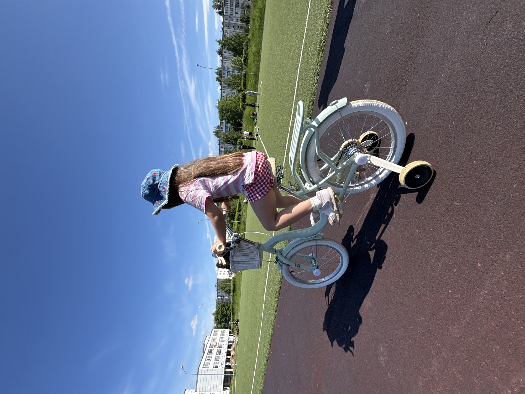 A young girl, viewed from the rear, rides a light mint-green bicycle equipped with white tires, white fenders, and functional training wheels on a reddish-brown asphalt track under a bright, clear blue sky with minimal cirrus clouds. The girl has long, straight brown hair and wears a blue denim bucket hat adorned with small red embroidered motifs, a pink and white striped short-sleeved top, red and white checkered shorts, and white sneakers. A white woven basket on the bicycle's handlebars contains a brown object. Strong sunlight casts distinct shadows of the bicycle and rider onto the track. To the left, a large, multi-story white building, possibly a school or public facility, stands adjacent to a green sports field with white demarcation lines; faint indications of ground-level construction or earthworks are visible near its base. To the right and rear, a row of multi-story, pre-fabricated residential buildings, typical of Soviet-era architecture in Chaikovsky, Russia, is partially visible beyond a line of trees. Several indistinct human figures are present on the distant green field, engaged in unidentifiable activities. The overall environment depicts an outdoor recreational area within an urban setting in the city of Chaikovsky, Russia.
