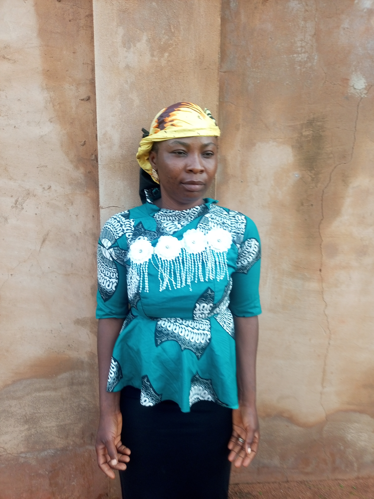 A woman is standing in front of a textured, light brown wall. She is wearing a teal peplum top with a black and white patterned print and white floral embellishments with white fringe. She also wears a black skirt and a yellow headscarf with an orange and brown pattern. Her left hand is visible and has rings on two fingers. The image appears to be taken outdoors. The location is identified as Tempure, Nigeria.