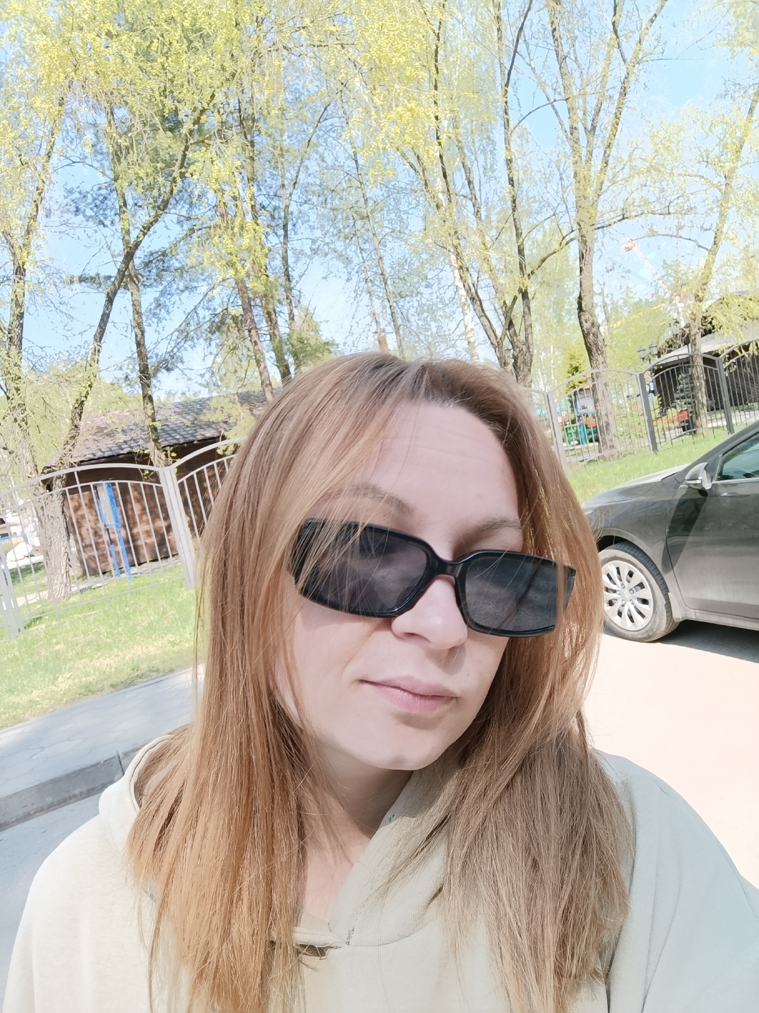 The image shows a woman with light brown, shoulder-length hair and wearing black rectangular sunglasses. She is wearing a light-colored hoodie. Behind her is a scene with trees in the background, a low wooden building with a fenced enclosure to the left, and a dark-colored car on the right. The setting appears to be an outdoor area near buildings and vegetation. The image was captured in the city of Chaikovsky, Russia.