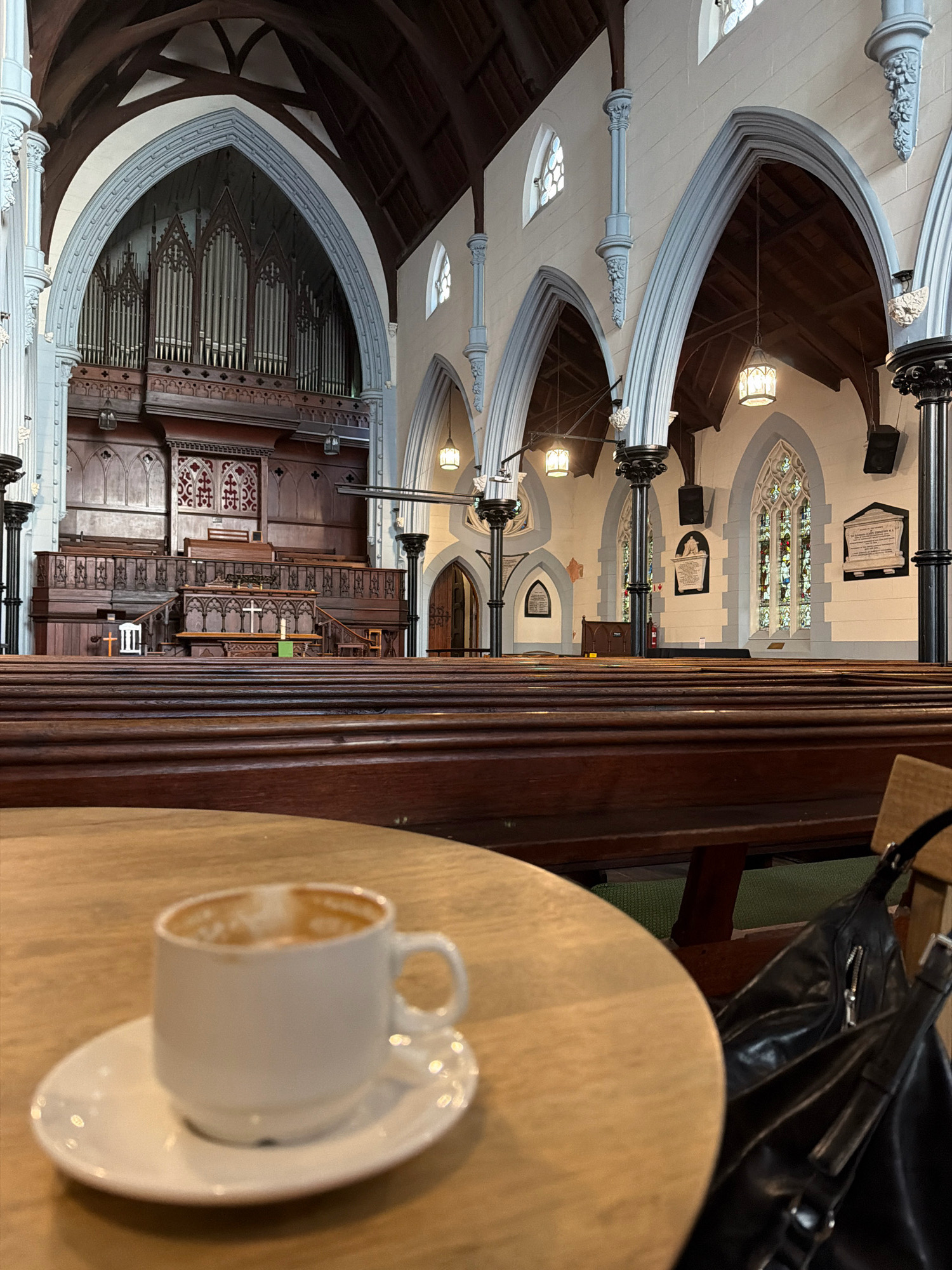 The image shows the interior of a church in Cape Town, South Africa. In the foreground, a partially consumed coffee sits in a white cup and saucer on a light-colored wooden table. To the right of the table, a black leather bag rests on a wooden bench. The background reveals rows of dark wooden pews, characteristic of church seating. Further in the background, a large pipe organ is visible, with intricate woodwork and ornate details. The church features high vaulted ceilings, stained-glass windows, and arched doorways, indicative of Gothic architecture. Natural light streams in through the windows, illuminating the space. The overall atmosphere is one of quiet contemplation and historical significance.