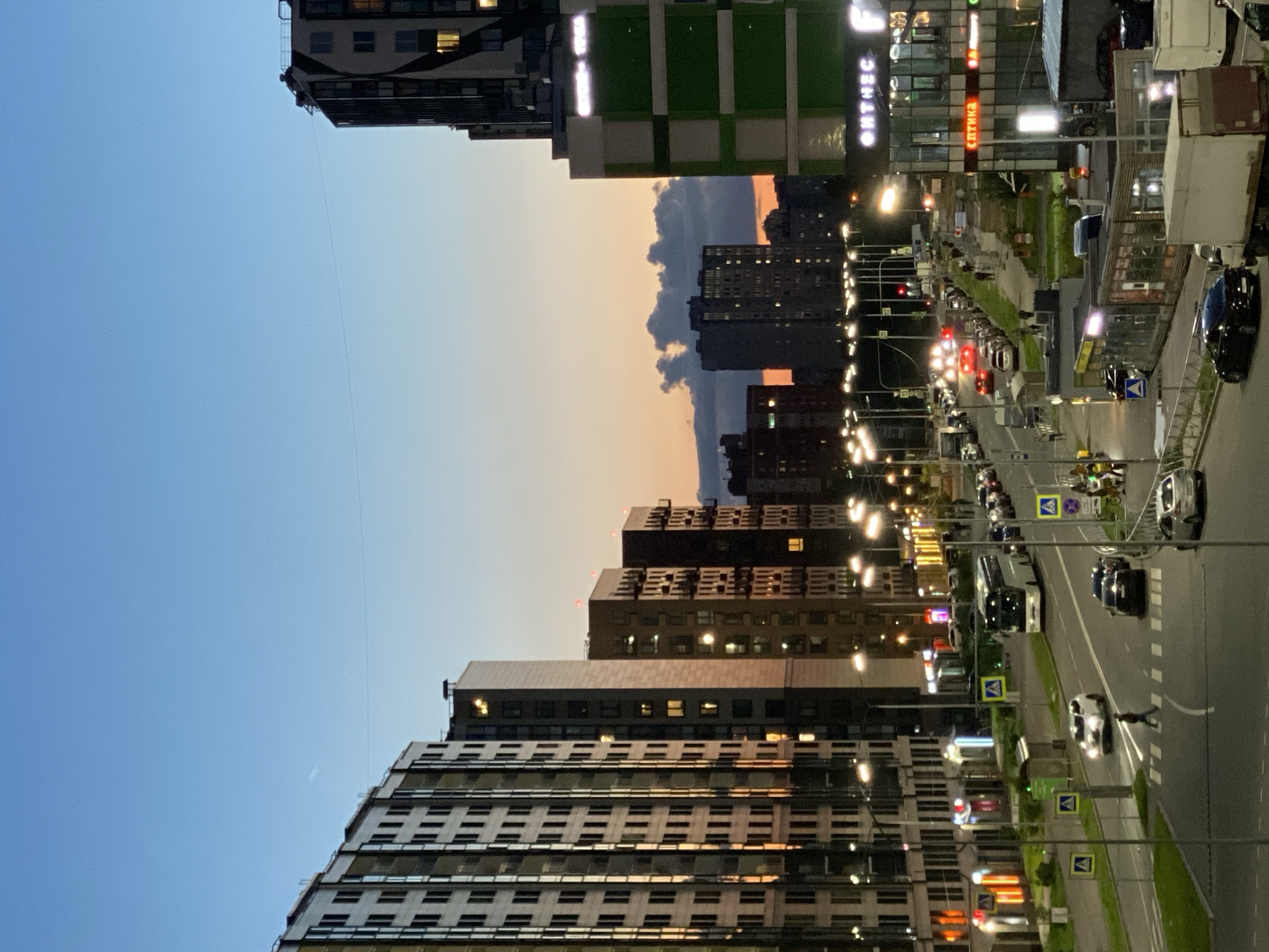 The image depicts a city street scene at dusk in Imeni Sverdlova, Russia. Tall residential buildings with illuminated windows line both sides of a multi-lane road. Several cars and a bus are visible on the road, with streetlights illuminating the scene. Pedestrians are crossing the street at a designated crosswalk. In the background, the sky displays a gradient of blue and orange hues, with clouds illuminated by the setting sun. Commercial buildings with illuminated signage are also present, including one with the word "ОНТНЕС".