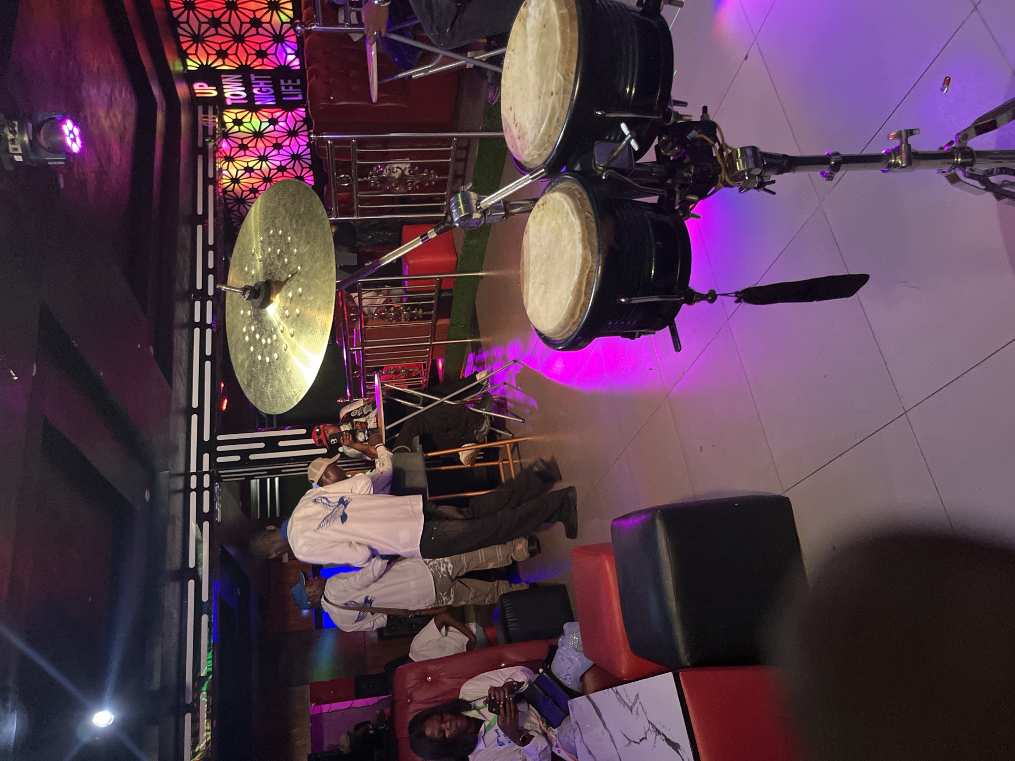 The image depicts the interior of a nightclub or bar. In the foreground, a set of bongo drums on a stand are prominently featured, illuminated by purple lighting. A large cymbal is positioned above the drums. To the left, a woman is seated on a red tufted chair, looking at her phone. She is holding a black purse on her lap and wearing a white top. In the background, several people are standing and interacting. One man in a white shirt with an eagle emblem and a blue baseball cap is facing away from the camera. Another man in a similar white shirt is standing beside him. Further back, a man in a white shirt and a baseball cap is seated at a high table, and another person in a dark shirt is visible. The environment is dimly lit with vibrant neon lights in shades of pink, purple, and blue illuminating the space. A decorative wall with a geometric pattern and neon signs reading "UP TOWN NIGHT LIFE" is visible in the background. The floor is tiled. The overall atmosphere suggests a lively evening entertainment venue.