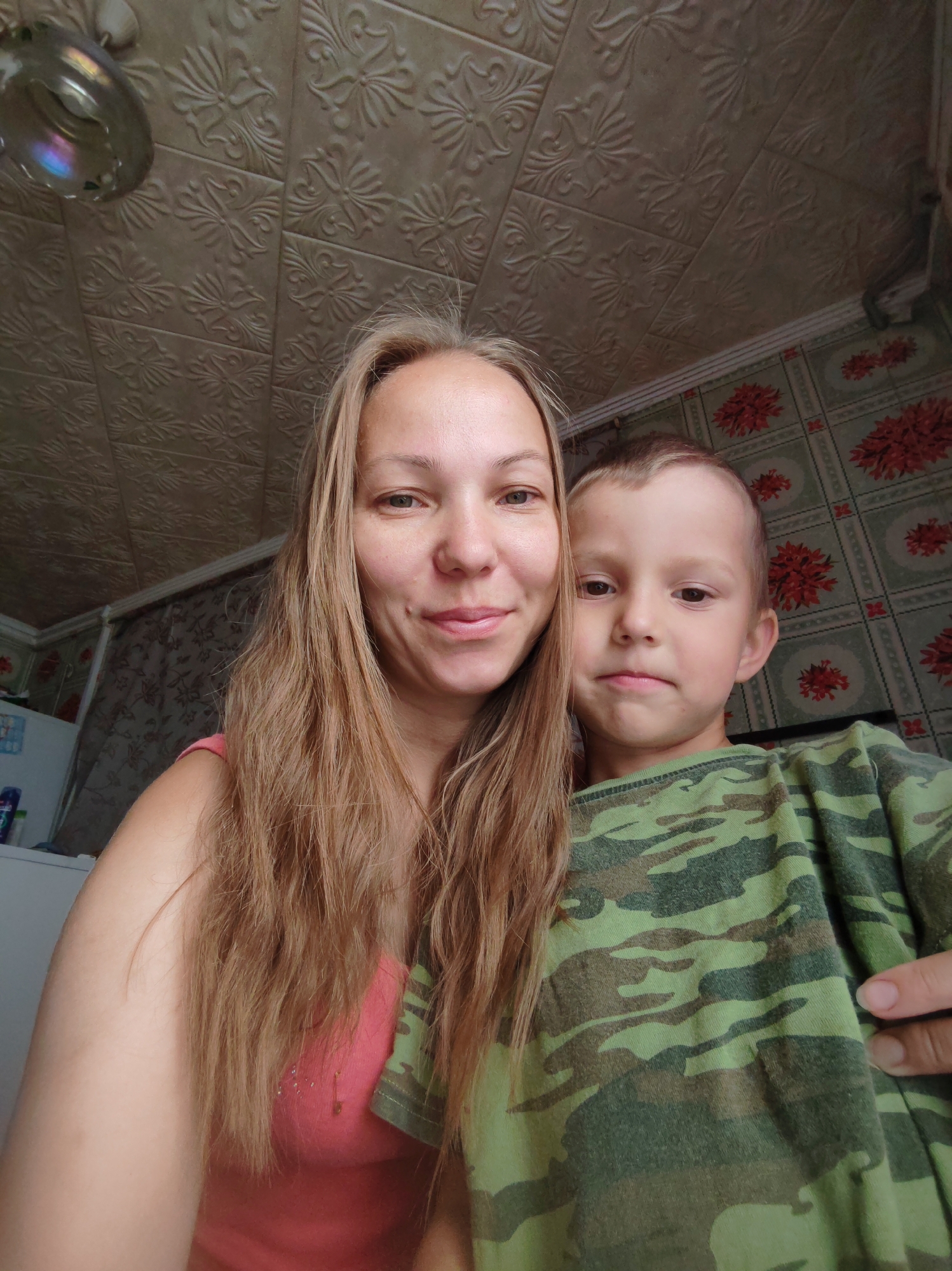 A medium shot photograph captures a woman and a young boy posing for a selfie indoors, likely in a bathroom, judging by the ceiling texture and shower head visible in the upper left corner. The woman, on the left, has long, blonde hair and is wearing a pink tank top. The boy, on the right, is wearing a green camouflage t-shirt. They are both looking towards the camera. The background features patterned wallpaper and a white refrigerator. The overall lighting suggests natural light. The location is specified as Чухлэм, Russia.