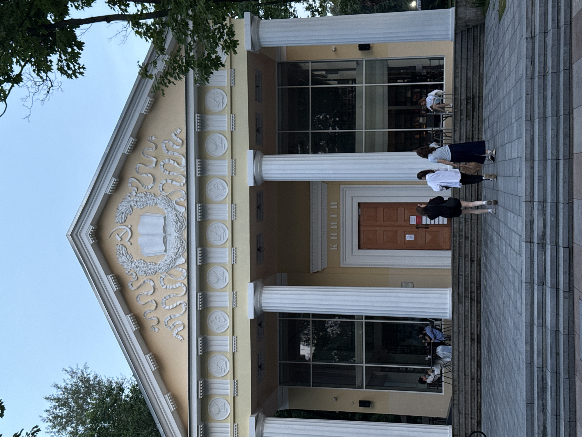 The image shows the exterior of a building with classical architectural elements, located in Moscow, Russia. The building is painted yellow and features white columns and decorative friezes. Above the entrance, there is a pediment with a bas-relief depicting a hammer and sickle, an open book, and a laurel wreath, indicating a Soviet-era aesthetic. The word "КНИГИ" (Books) is visible above the central doorway.
In front of the building, a wide set of stone stairs leads up to a portico with large glass windows. Inside the building, through the windows, bookshelves are visible, suggesting it is a library or a bookstore. Several people are seated at tables outside the building on the right side, engaged in conversation.
On the left side of the stairs, three young women are walking towards the entrance. One woman in the front is wearing a black jacket and shorts, the middle woman wears a white shirt and black pants, and the woman behind her is in a blue skirt and white top. On the right side, near the entrance, another person is seated outside. The surrounding area appears to be a public space with trees visible in the background and a cloudy sky above. The overall impression is of a well-preserved historical building in an urban setting.