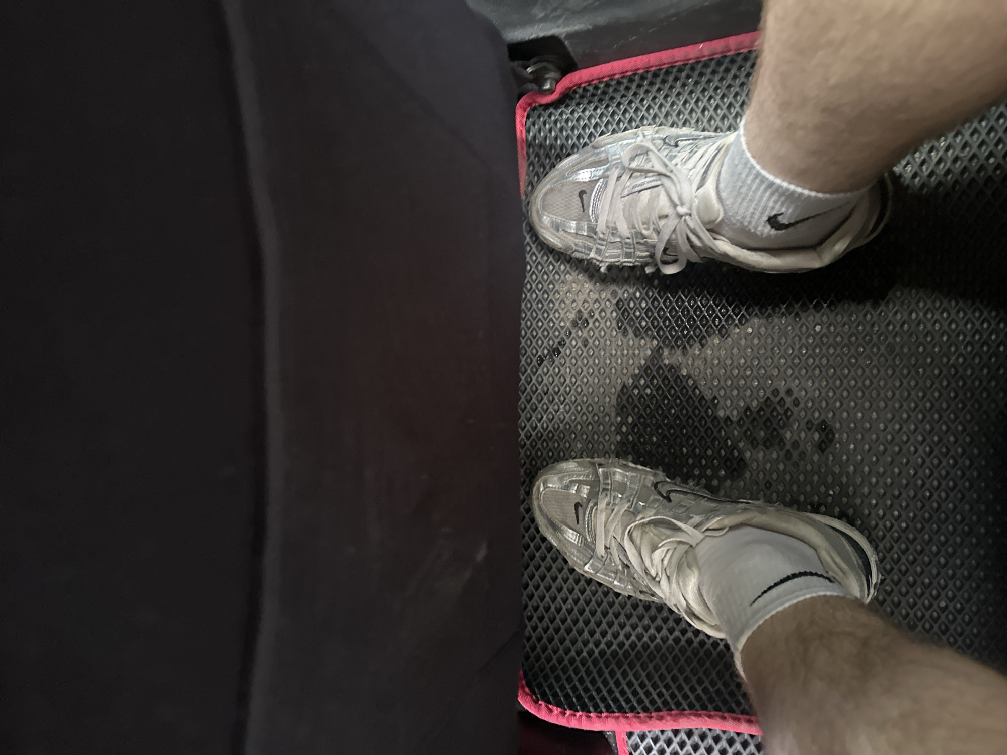 The image shows a first-person perspective of two feet wearing athletic shoes and socks, resting on a black floor mat with a red trim. The mat is textured with a diamond pattern. The shoes are light gray with white laces and appear to be Nike brand, as indicated by the visible swoosh logo on the side and on the socks. The background is a dark, textured surface, likely the back of a car seat. The lighting is dim, suggesting an indoor environment or a dark setting like the interior of a vehicle. There is no visible indication of the location in Birobidzhan, Russia.