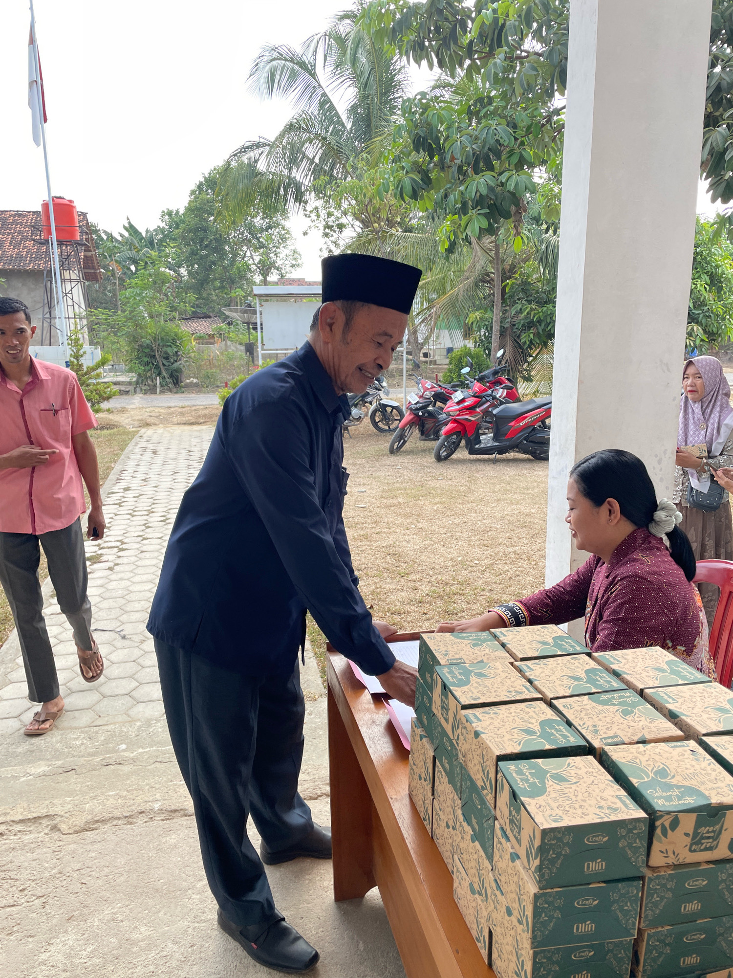 In Kagungan Ratu, Indonesia, an outdoor scene depicts an exchange at a wooden table laden with stacked boxes. Two men and two women are present. The man in the foreground, wearing a dark blue shirt, black peci, and black pants, appears to be handing a document to a woman seated at the table. The woman, dressed in a maroon garment with intricate patterns on the sleeves, wears her hair in a bun and smiles as she receives the item. To her right, another woman in a light purple hijab and patterned dress observes the interaction. Behind the man in the blue shirt, another man in a pink collared shirt and grey pants walks away from the table, looking back. The environment features a paved walkway, dry grass, trees, and parked motorcycles in the background. The sky is overcast.