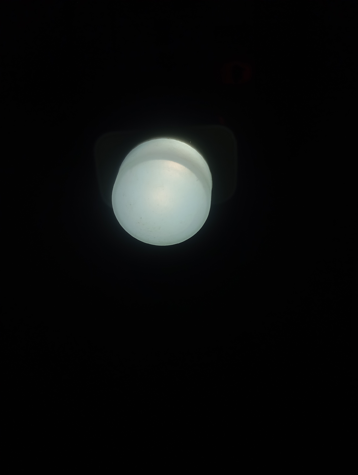 A single light bulb hangs from a dark ceiling in an otherwise black, empty room. The light bulb is round and frosted, emitting a soft white glow. The background is completely black, with no discernible features or details. The image is taken from a low angle, looking up at the light bulb.