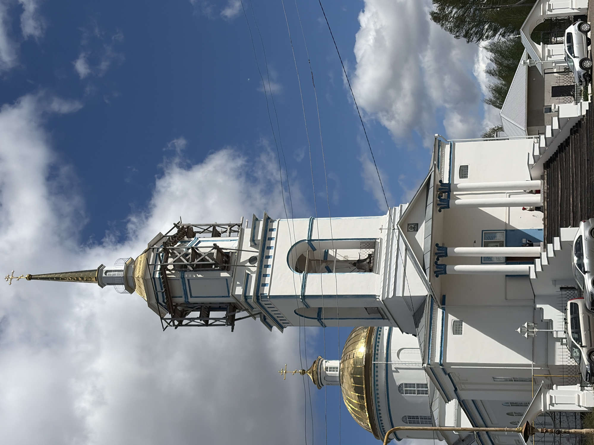The image depicts a white and blue Russian Orthodox church in Mozhga, Russia. The church features golden domes and is partially covered in scaffolding, indicating ongoing construction or renovation. Power lines are visible against a cloudy sky. A building is adjacent to the church with cars parked next to it, with what appears to be stairs leading to the top.